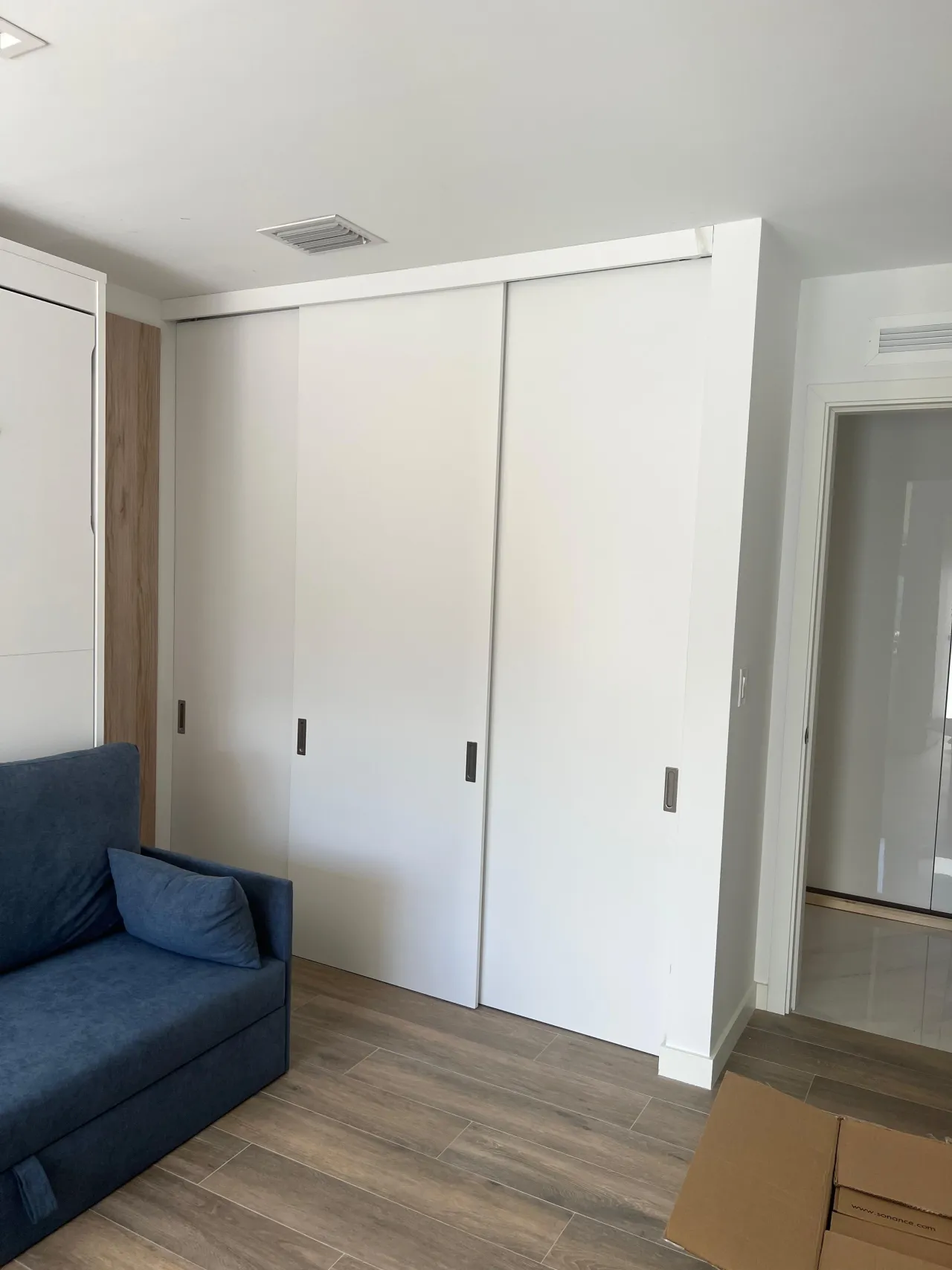 White sliding closet doors with small metal tab handles in a modern living room, next to a blue upholstered sofa and wood-look floor