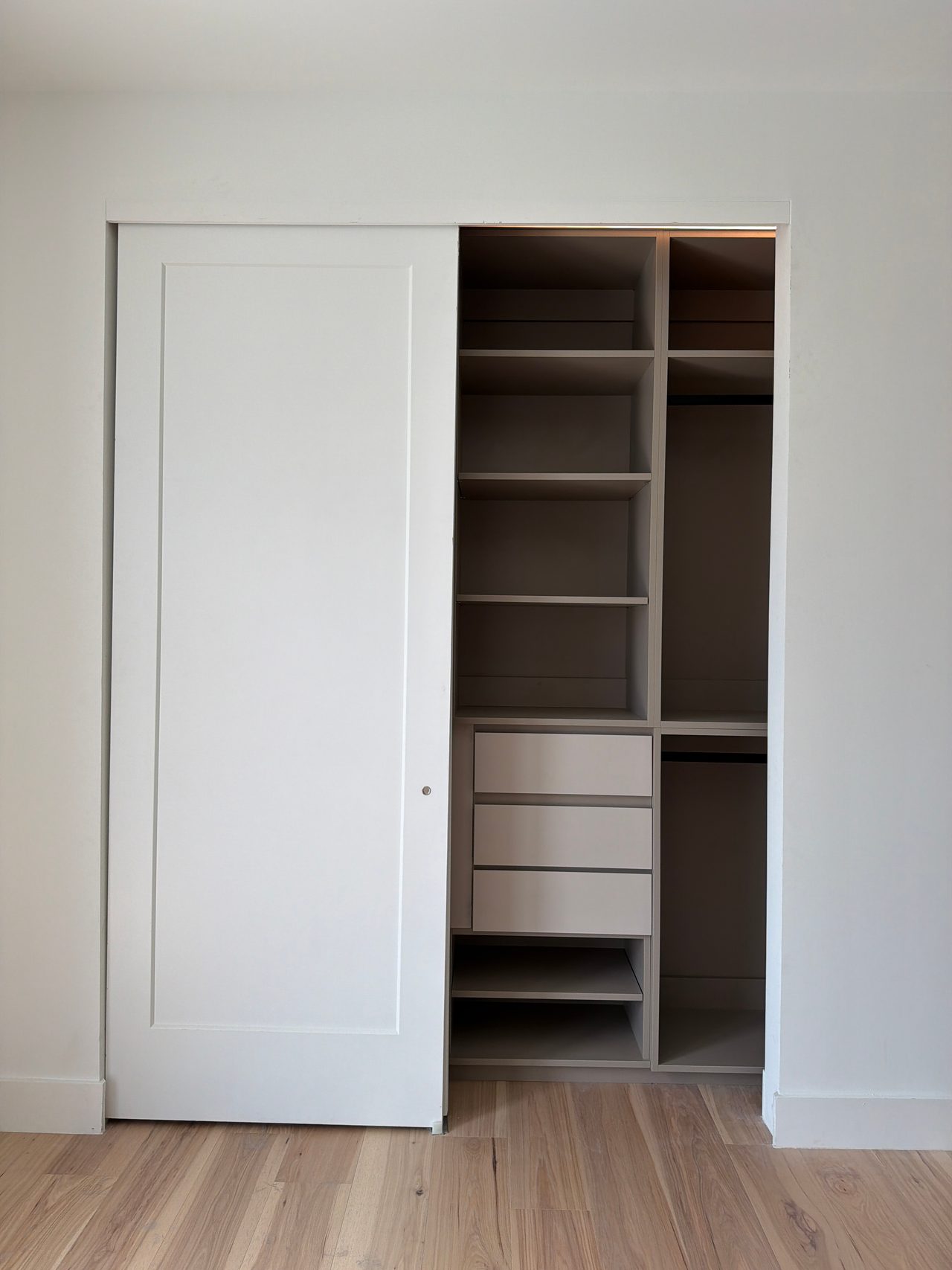 Closet with a white sliding panel door pushed open to show greige shelving, three drawers and hanging rods, over a light wood-grain floor