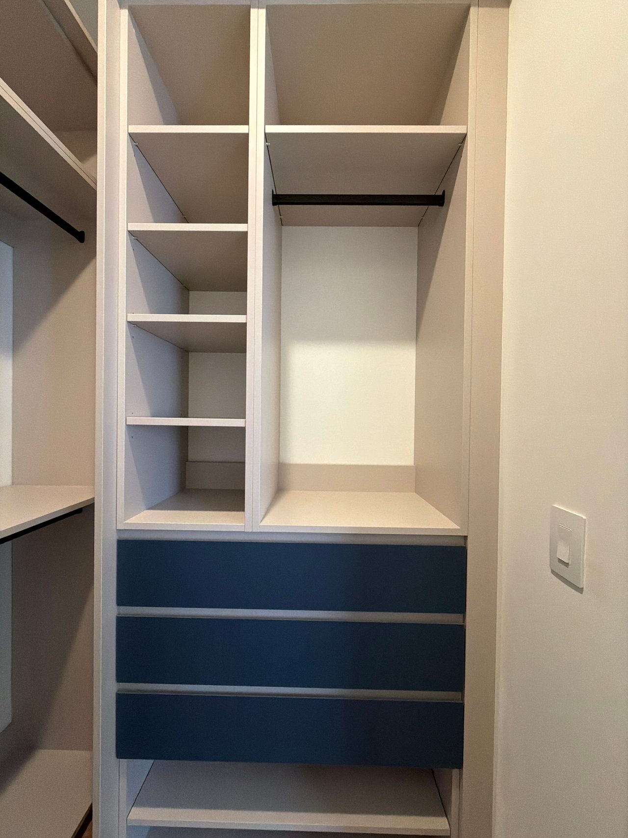 White closet unit with open shelf compartments, a black hanging rod and three navy blue drawers below, beside a wall light switch