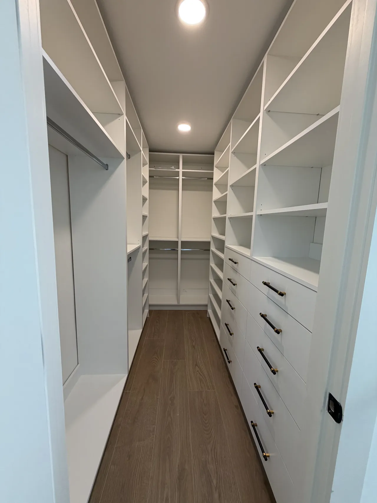 White walk-in closet corridor with open shelving on both sides, a hanging rod on the left, and a white drawer stack with dark handles