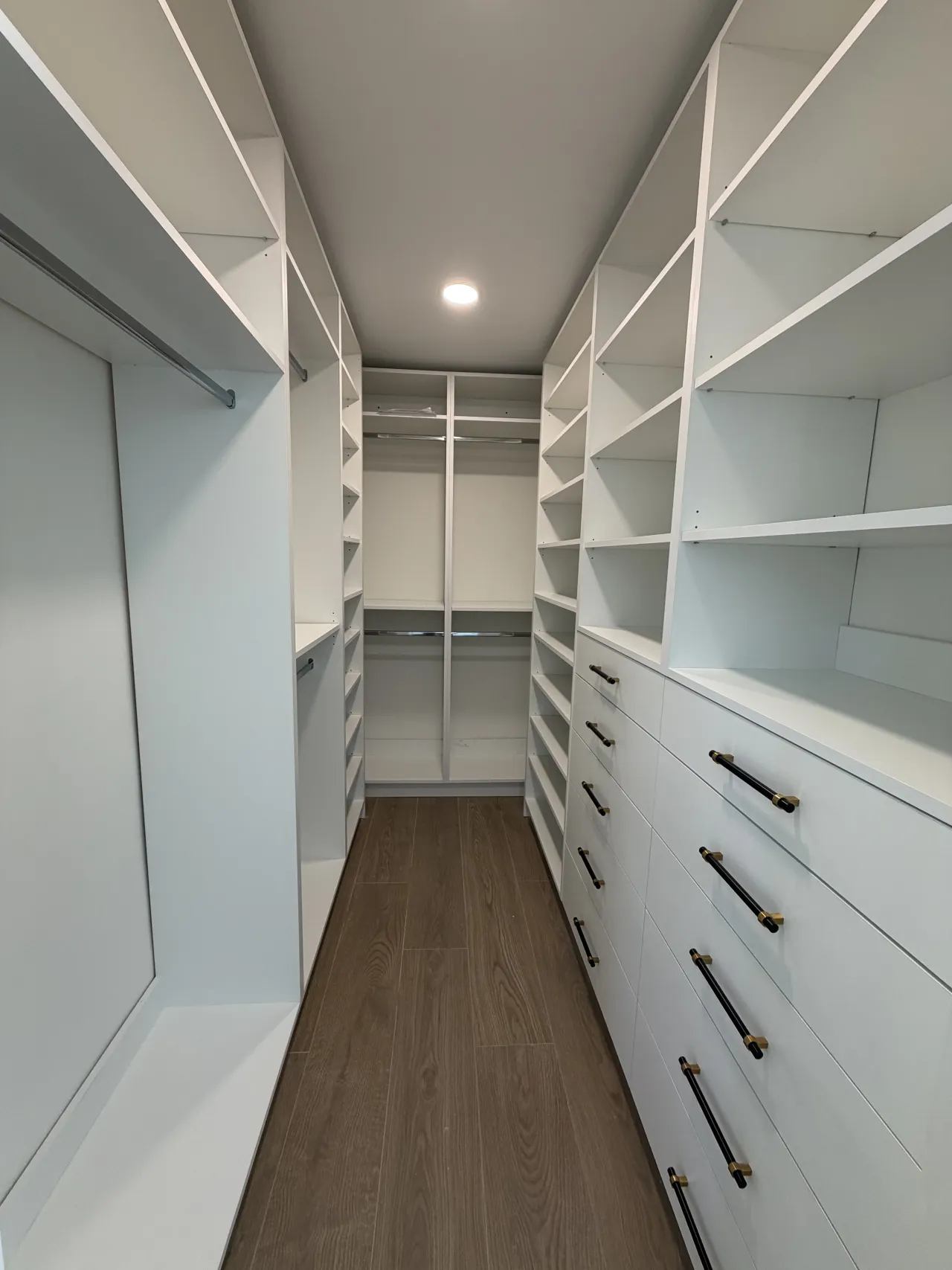 White walk-in closet corridor with open shelving on both sides, hanging rods, and a dark-handled drawer bank on the right, dark wood-look floor