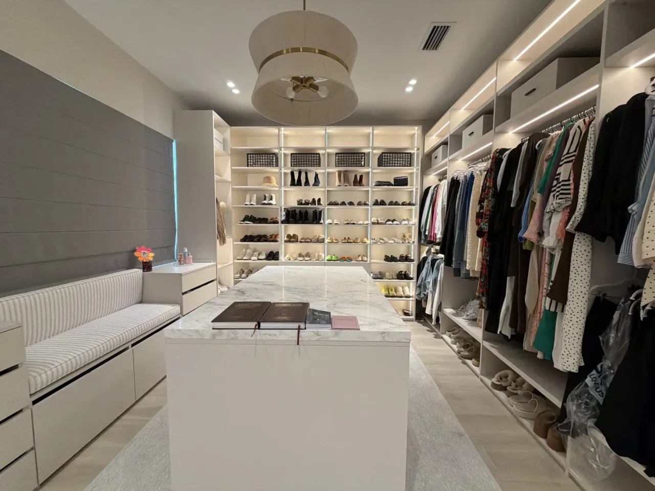 White walk-in closet with a stone-top island, open shoe shelving with LED lighting, hanging clothes rods, and an upholstered bench