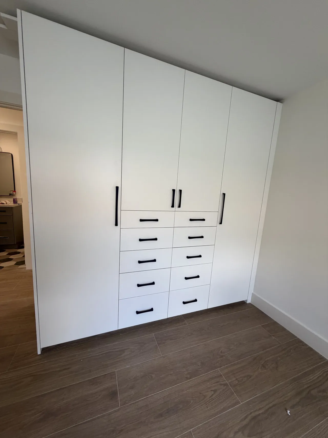 White wardrobe with four tall doors and two center drawer columns with black bar pulls, seen at an angle beside an open doorway, dark wood-look floor