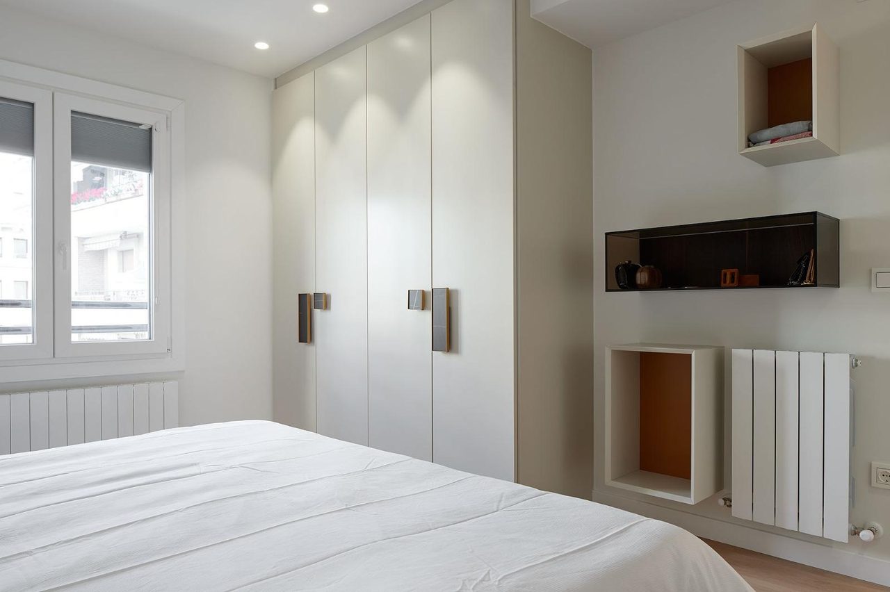 White wardrobe wall with dark handles beside a window, an orange-lined display cubby and a dark glass shelf display box, white radiator below