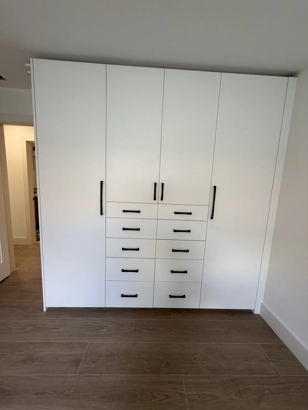 White wardrobe wall with four tall doors, two center columns of five drawers each, black bar pulls, an open doorway at left and dark wood-look floor