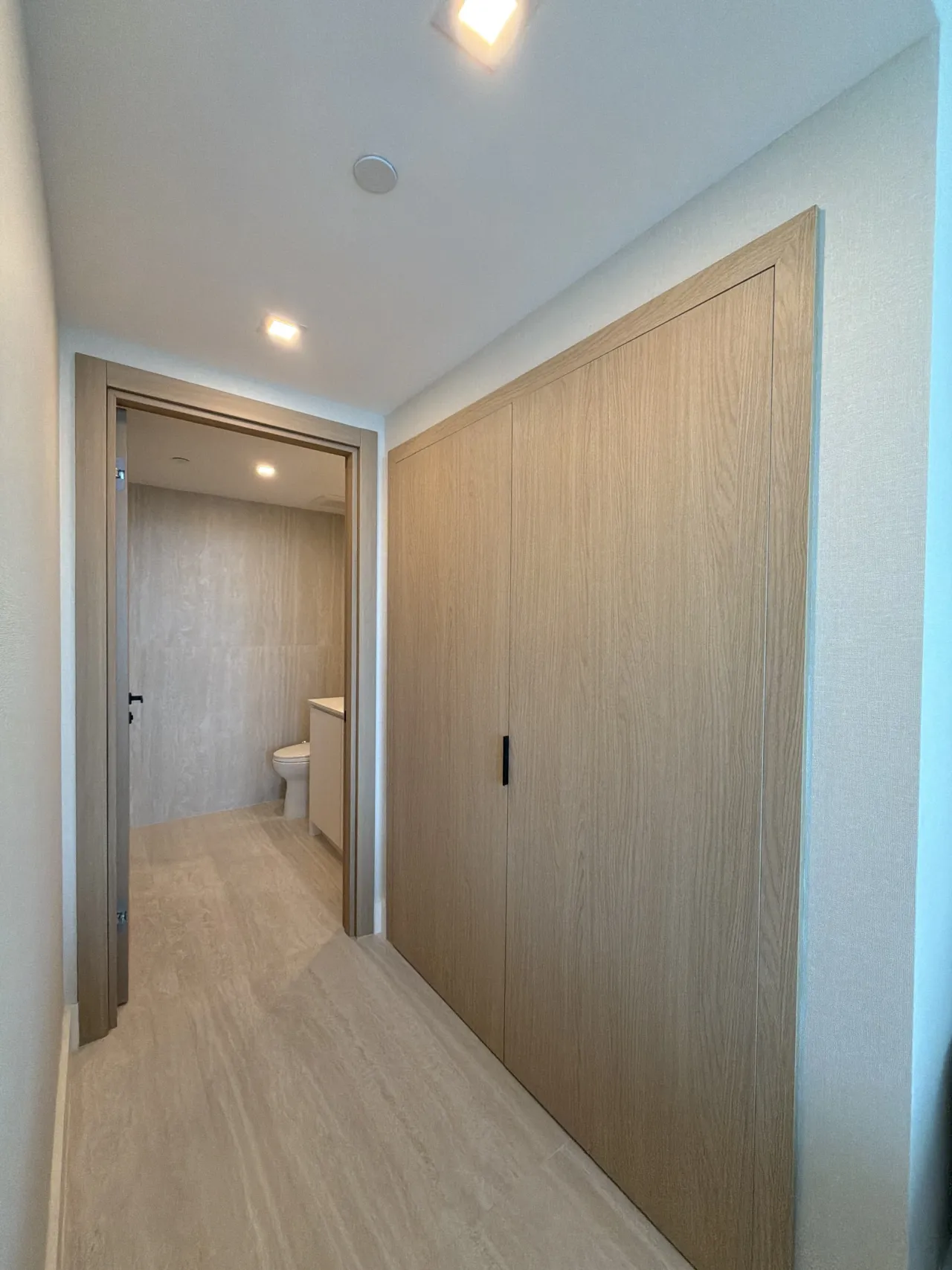 Wood-grain melamine panel closet doors in a hallway leading to a bathroom with a toilet visible, light stone tile floor