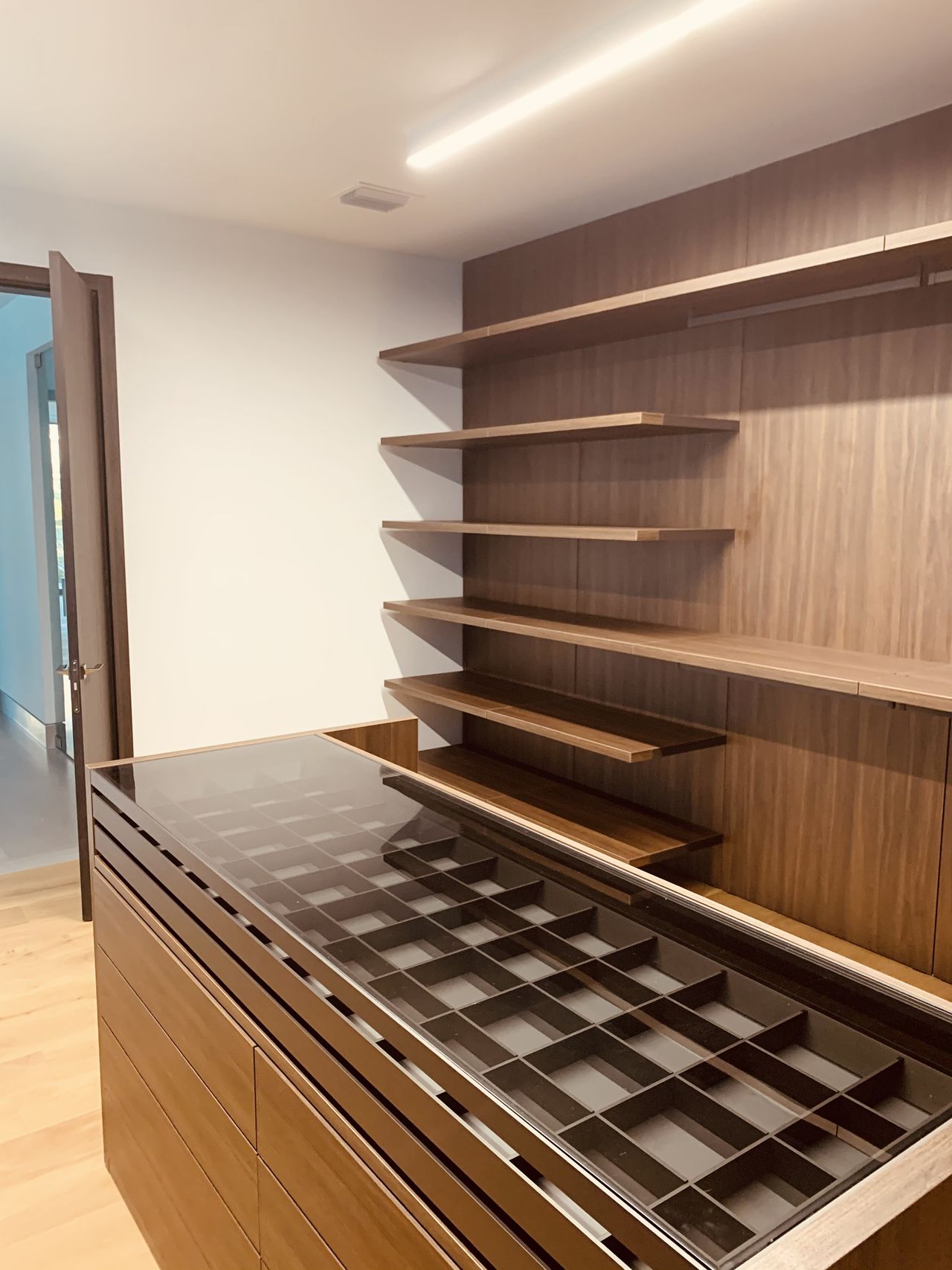Wood-grain walk-in closet with angled open shelving, a hanging rod, and an island with glass-topped divided compartment trays, light wood floor