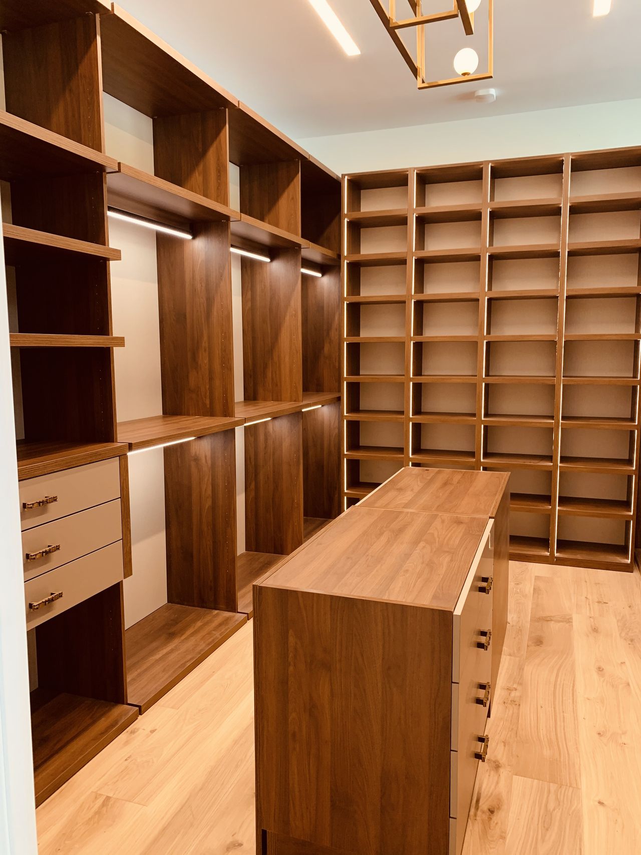 Wood-grain walk-in closet with lit hanging rods and a cream drawer bank at left, tall grid cubby shelving at right, gold-tone geometric pendant