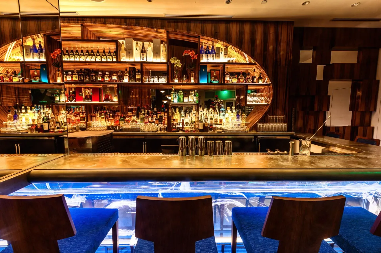 Bar counter with a backlit oval shelving arch of bottles, wood-grain paneling, and a blue-lit glass panel running straight below the metallic counter
