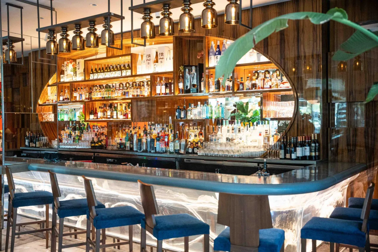 Bar with an oval wood-grain shelving arch of bottles, gold-tone glass pendants above, a metallic counter and blue upholstered barstools, leaf in foreground