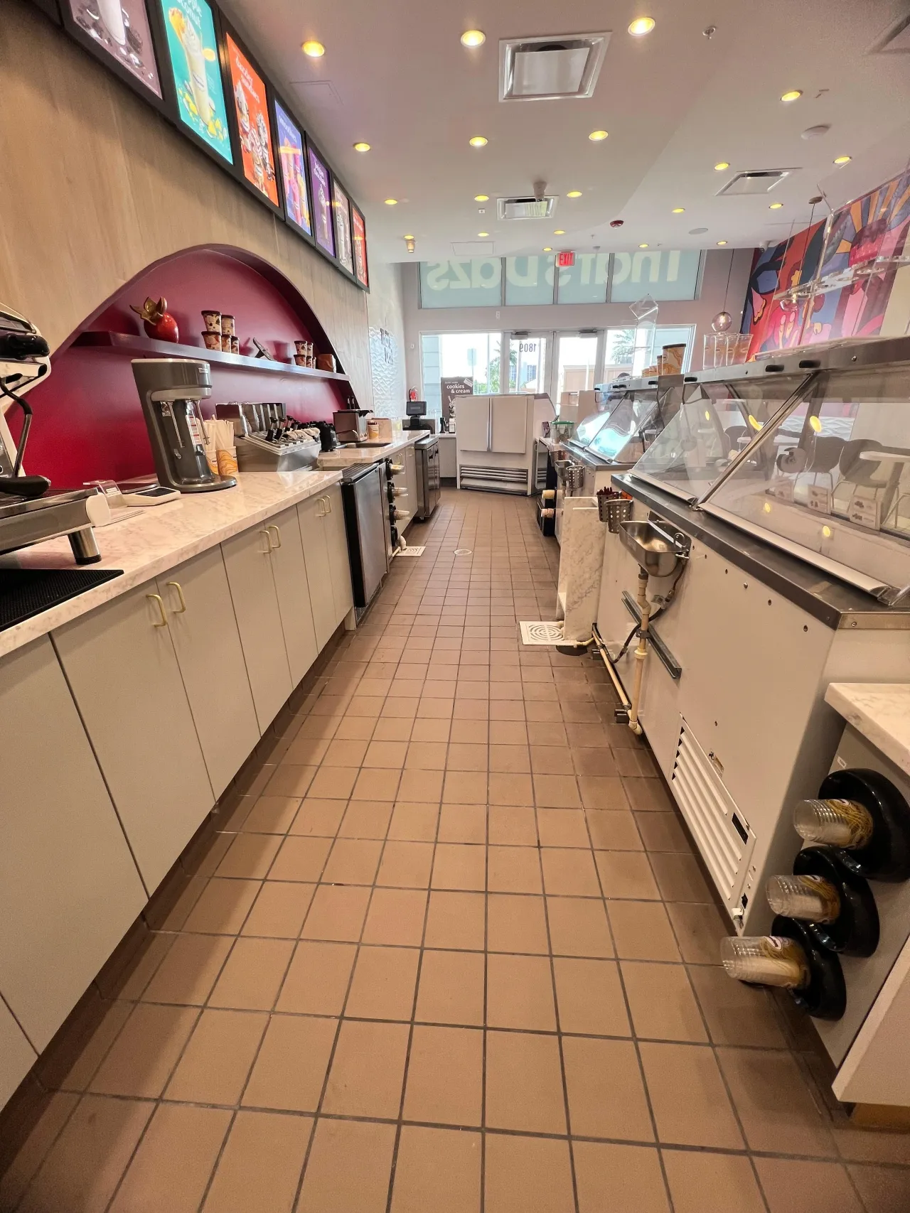Behind-counter prep aisle with tan tile floor, cream flat-panel cabinets with bar pulls, veined stone countertop and a dipping cabinet