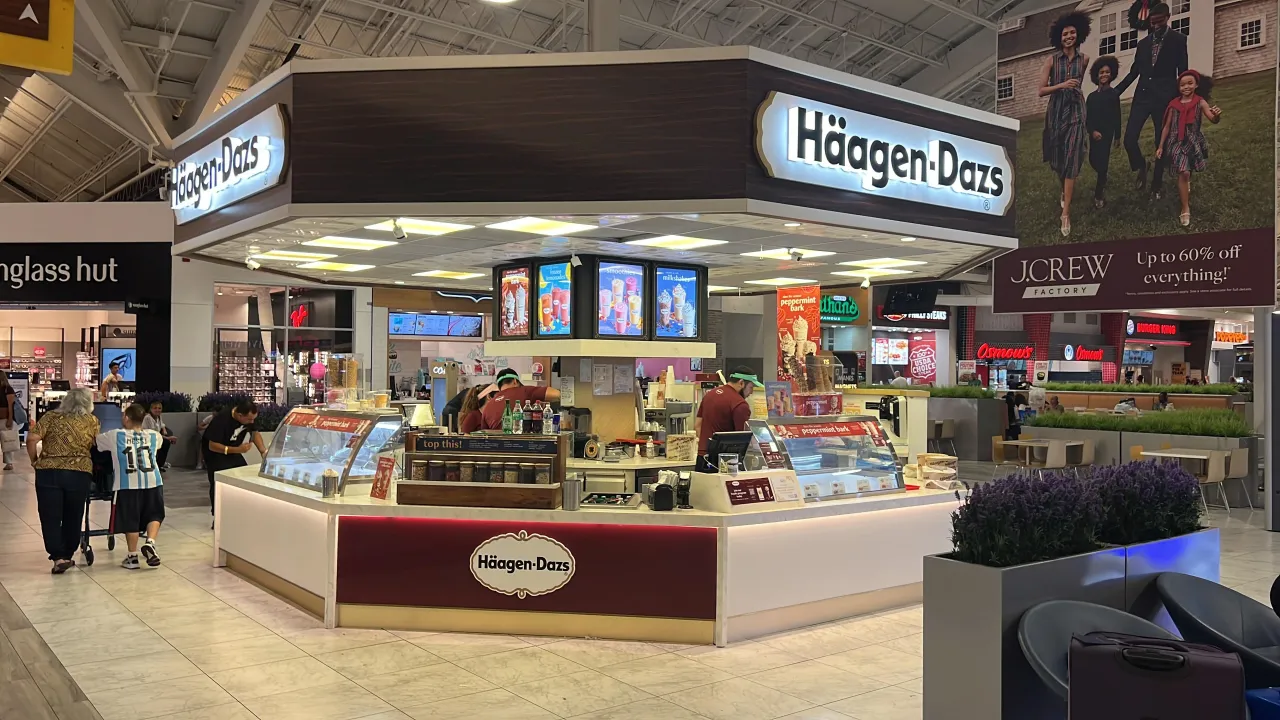 Corner mall kiosk with dark wood-grain fascia and canopy, illuminated white base, glass display case, located near a store corridor with shoppers