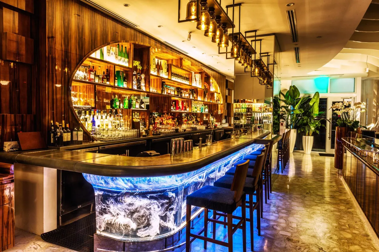 Curved bar front with a backlit blue glass base, wood-grain shelving stocked with bottles, and gold-tone glass pendant lights in metal frames above