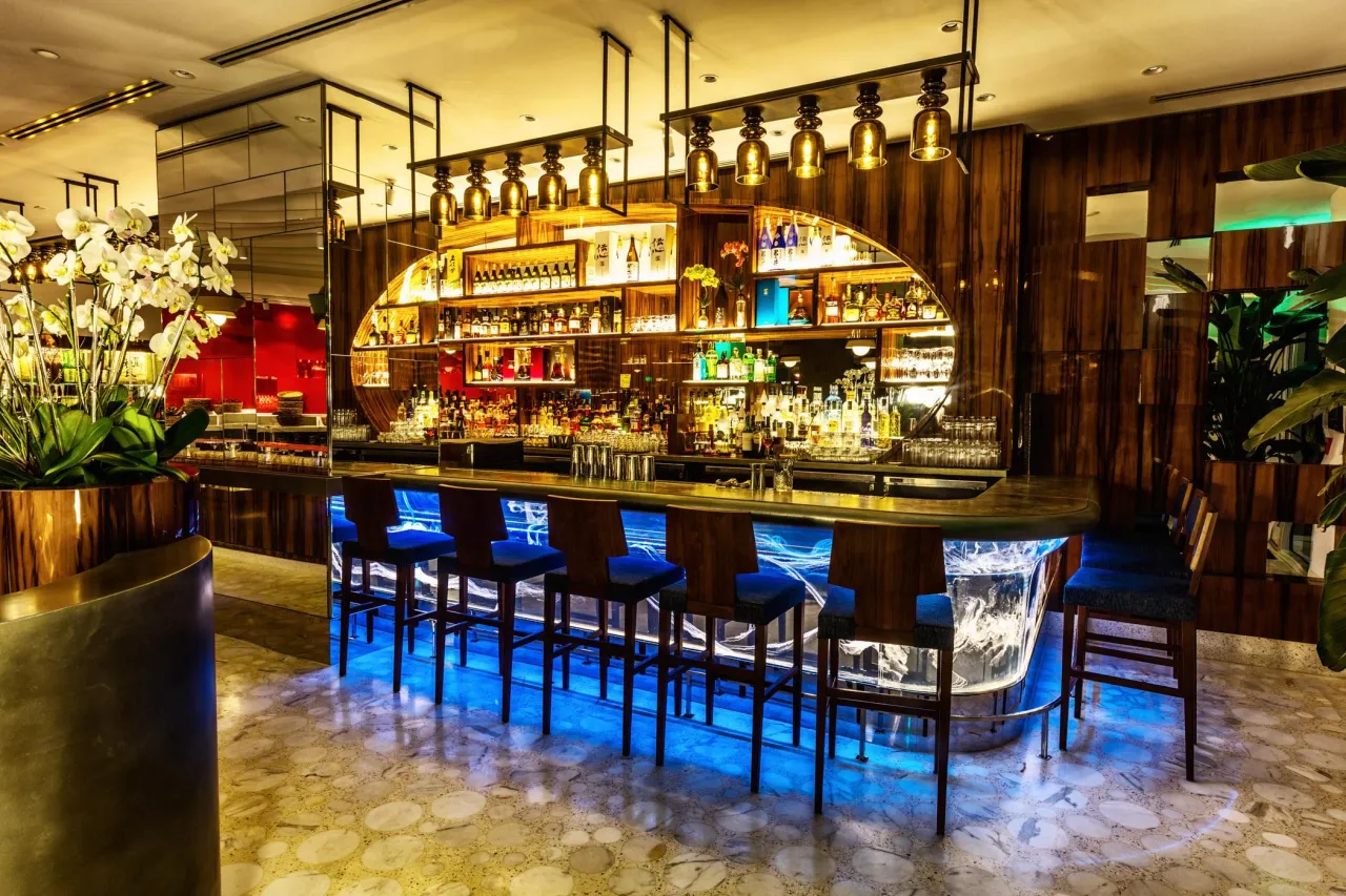 Curved restaurant bar with blue LED-lit glass base, dark wood millwork shelving stocked with bottles, gold-tone pendant lights above