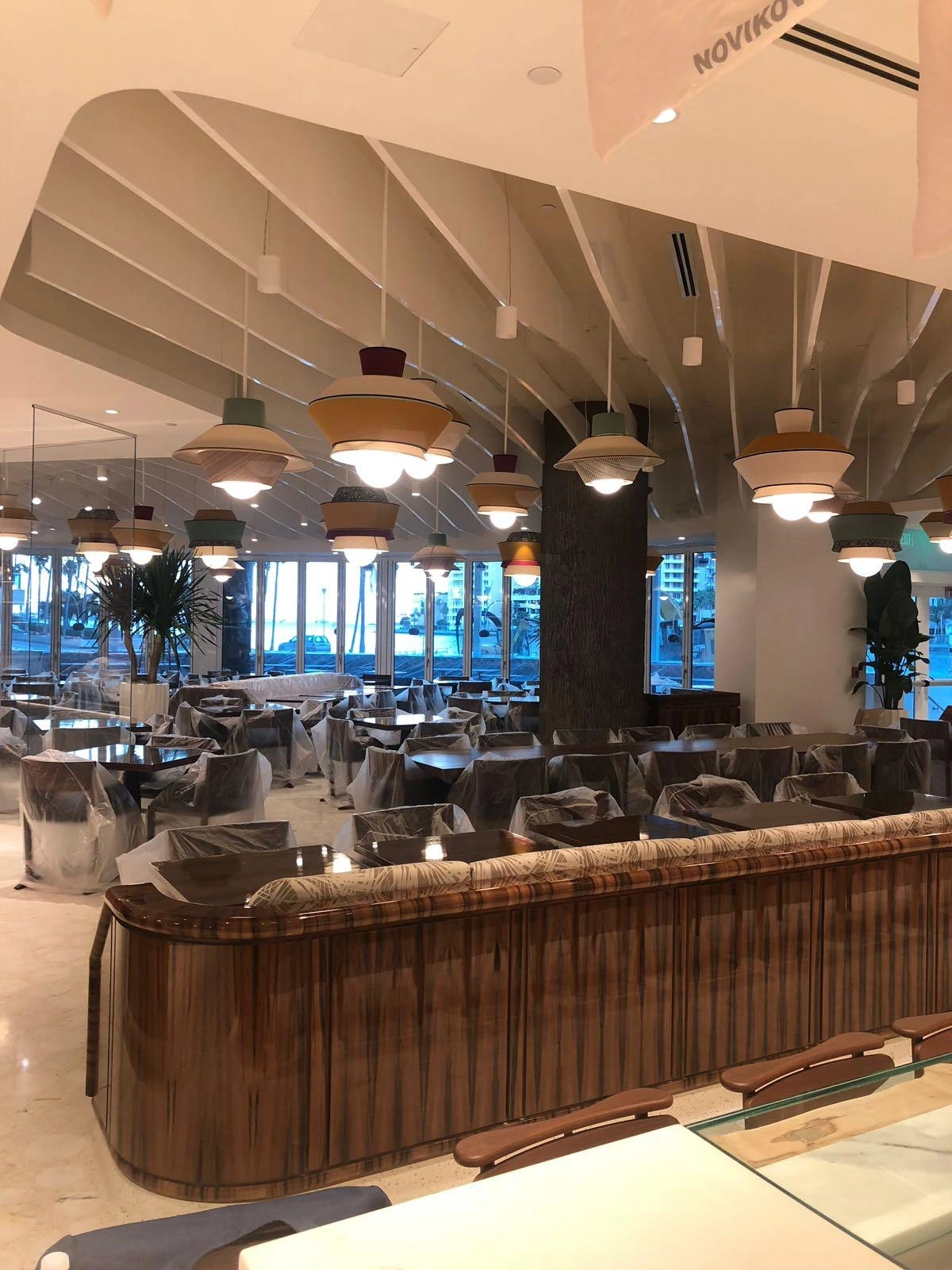 Curved wood-grain paneled divider with a patterned upholstered top rail, plastic-wrapped tables beyond, pendants under a white sculpted ceiling
