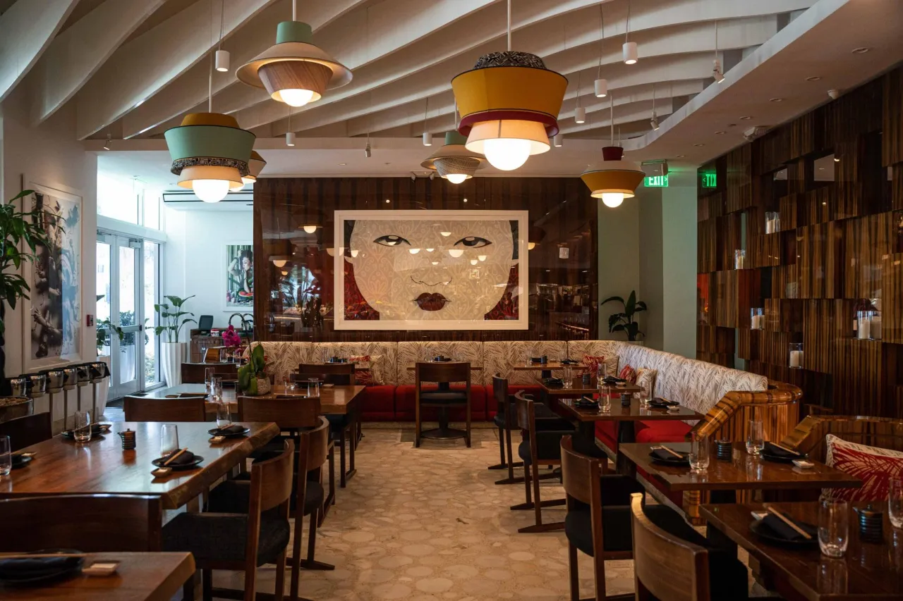 Dining area with a large framed face artwork on a wood-grain wall, long patterned banquette, wood-grain tables and colorful layered pendant lights