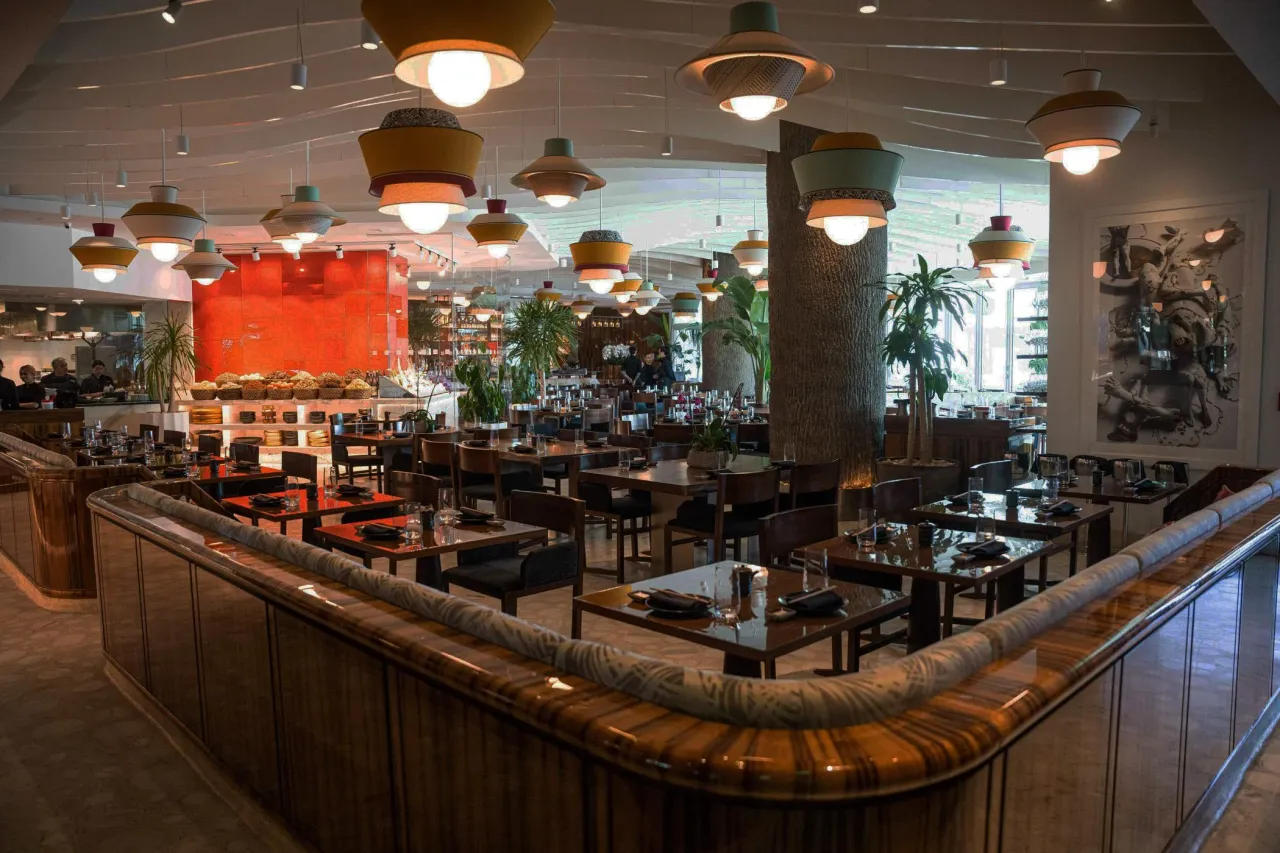 Dining room with a white sculpted wavy ceiling, colorful layered pendant lights, curved wood-grain paneled partition in front and a red glossy wall beyond