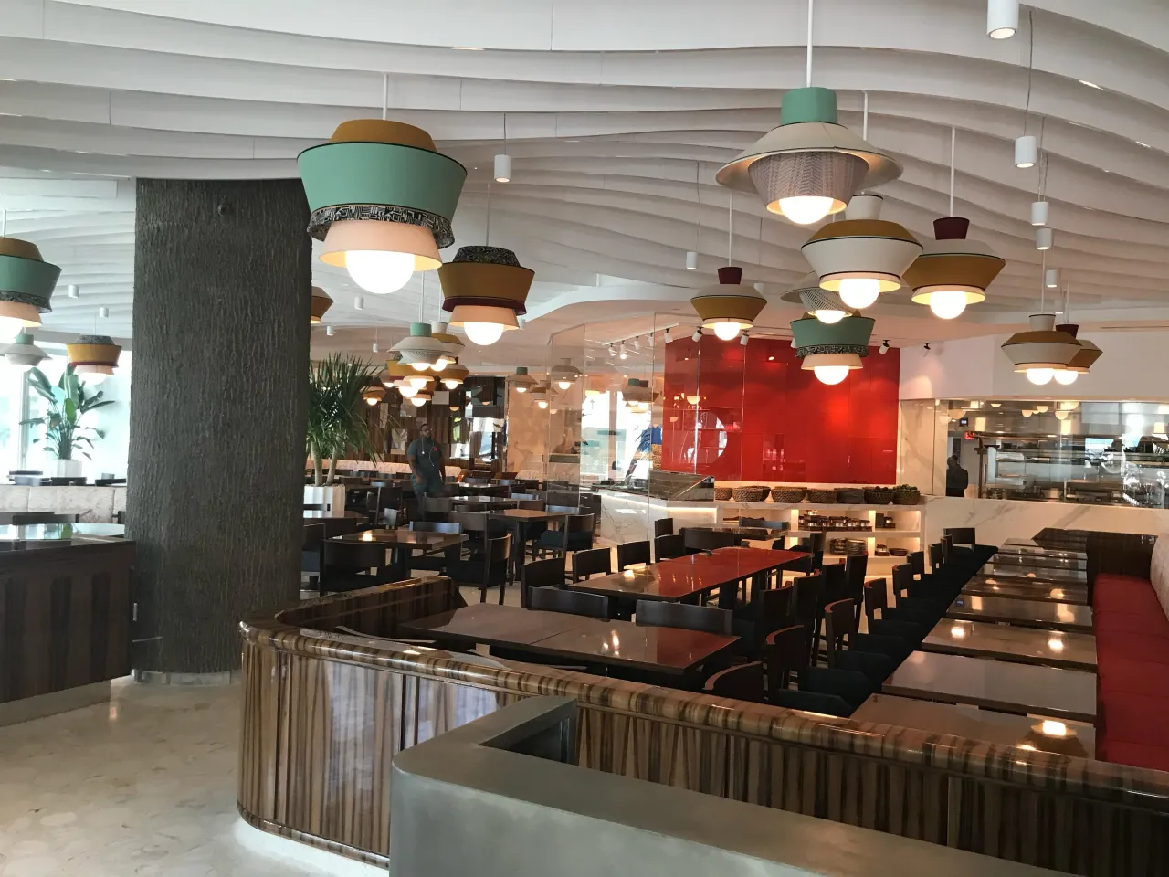 Dining room with a white sculpted wavy ceiling, rows of colorful layered pendant lights and a curved wood-grain paneled banquette in the foreground