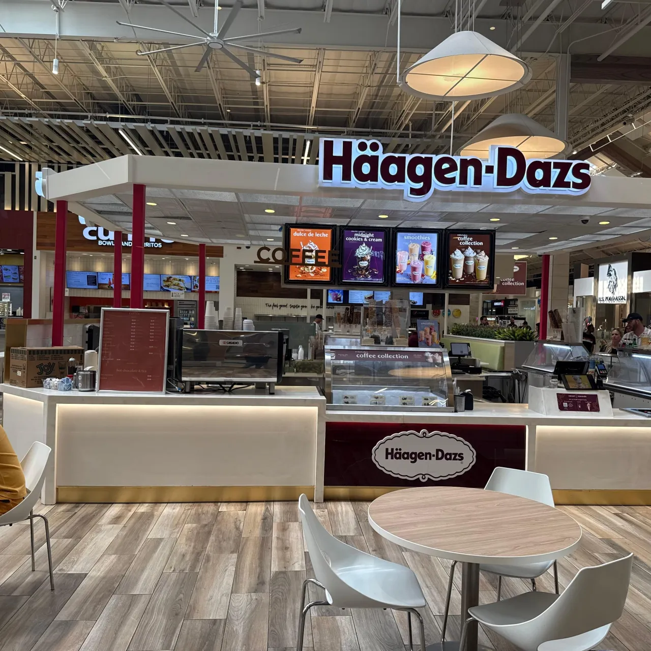 Food-court kiosk with an illuminated white counter base, dark red front panel, red posts and white canopy, glass display cases, wood-plank floor