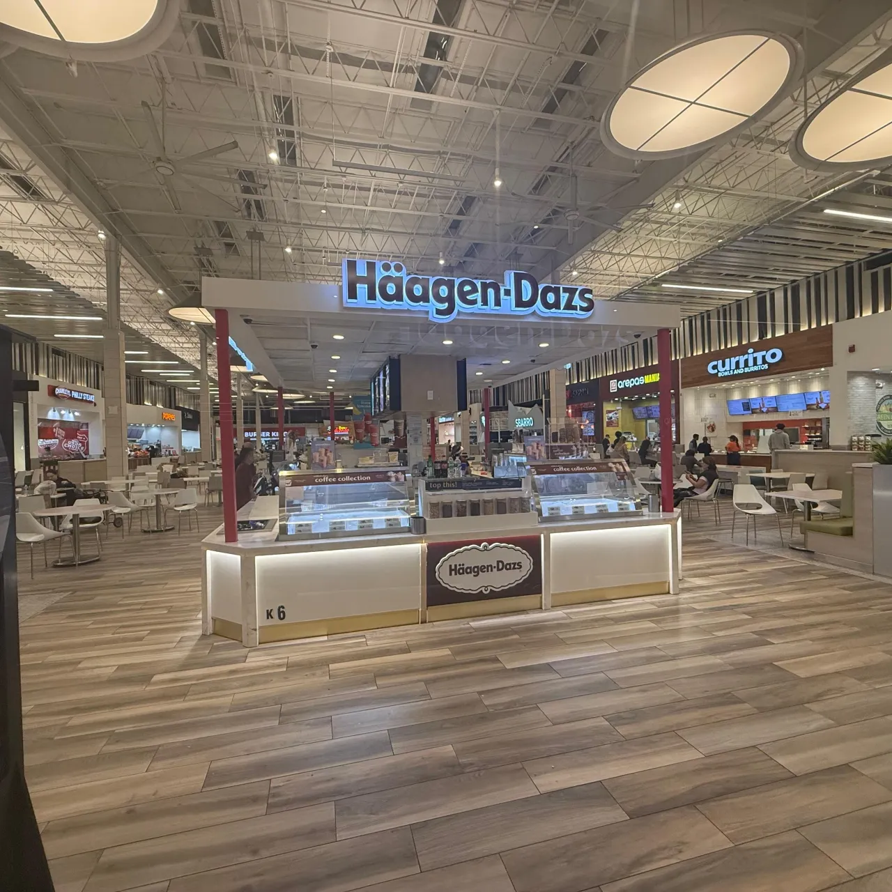 Island kiosk in a food court with an illuminated white base, dark red center panel, straight glass display cases on top, wood-look plank floor