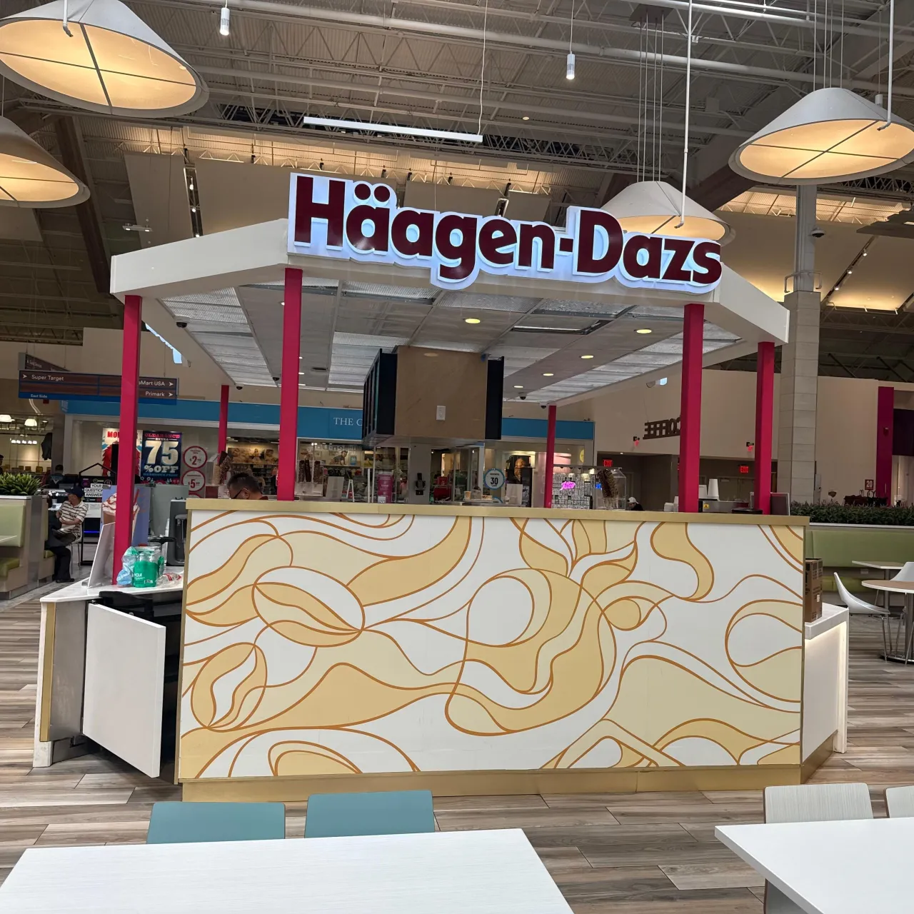 Kiosk counter clad in a cream panel with gold-tone swirl line pattern, red columns supporting a white canopy, mall seating around it