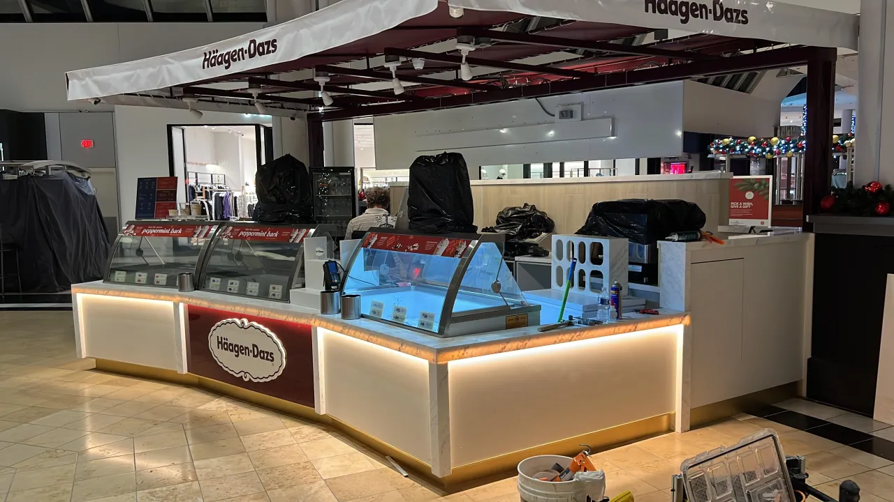 Kiosk under fit-out with veined stone counter top, glow-lit white base, dark red sign panel, glass display cases and a white awning canopy