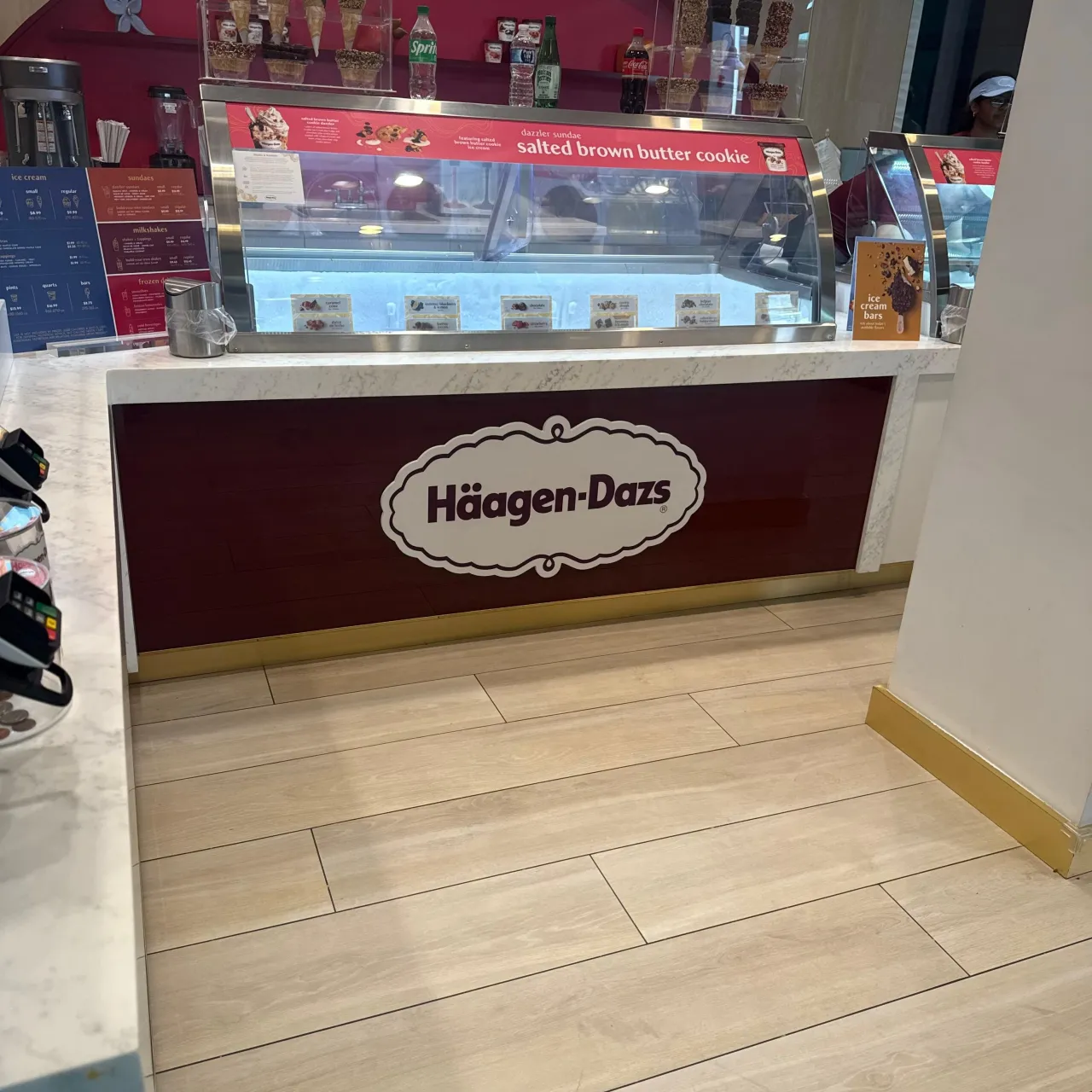 Maroon-fronted veined stone display counter with a curved glass case, menu strip above, drink bottles on shelf, light wood floor