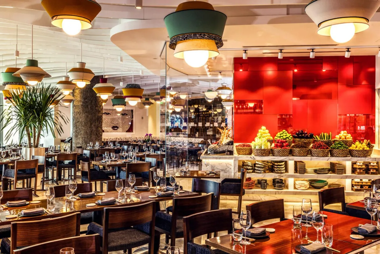 Open restaurant dining room with colorful drum pendant lights, a veined stone-fronted produce display counter, red glass wall behind