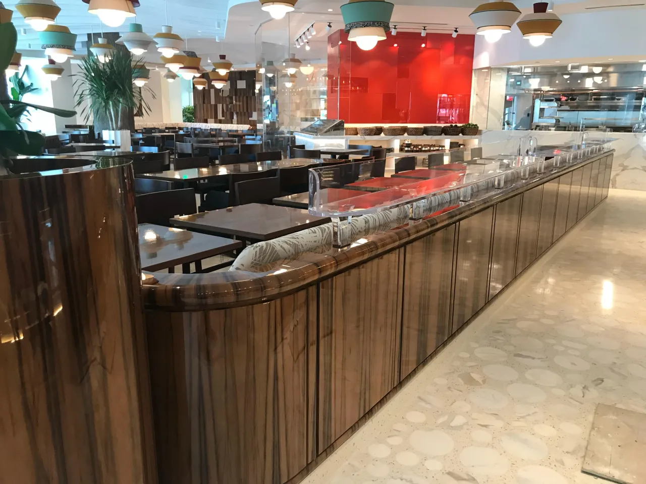 Restaurant dining room with a curved dark wood-paneled counter edge, veined stone server station, dining tables and colorful pendant lights