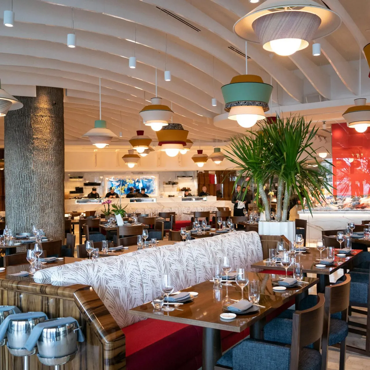 Restaurant dining room with a curved upholstered leaf-print banquette, wood dining tables, colorful pendant lights and a tall potted palm