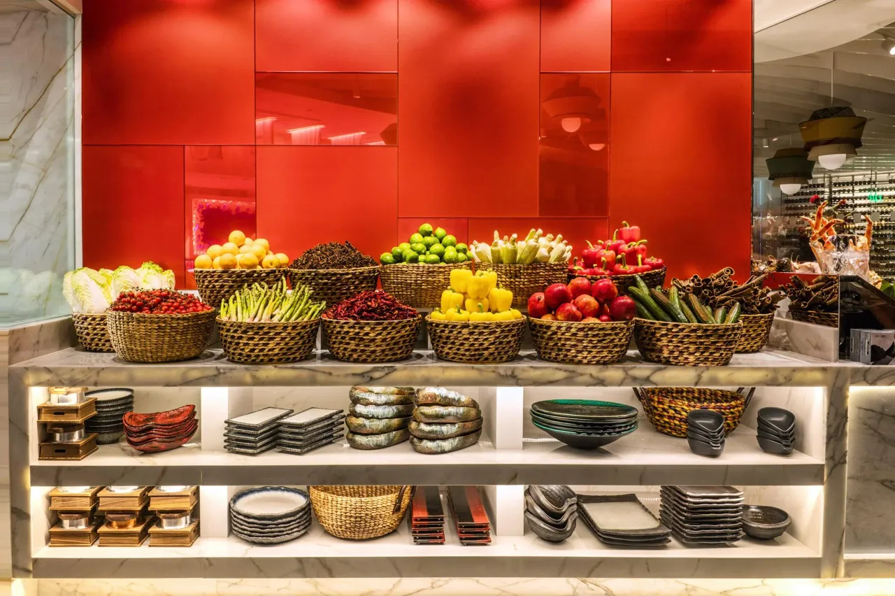 Restaurant fresh produce display station with woven baskets of vegetables and fruit atop a white veined stone counter, red glossy glass feature wall behind