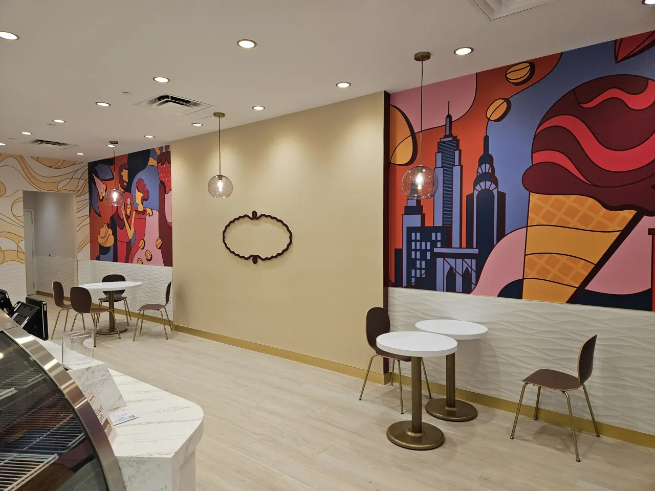 Seating nook with small round cafe tables and brown chairs against a cream wall with colorful ice-cream themed murals, veined stone counter edge at left