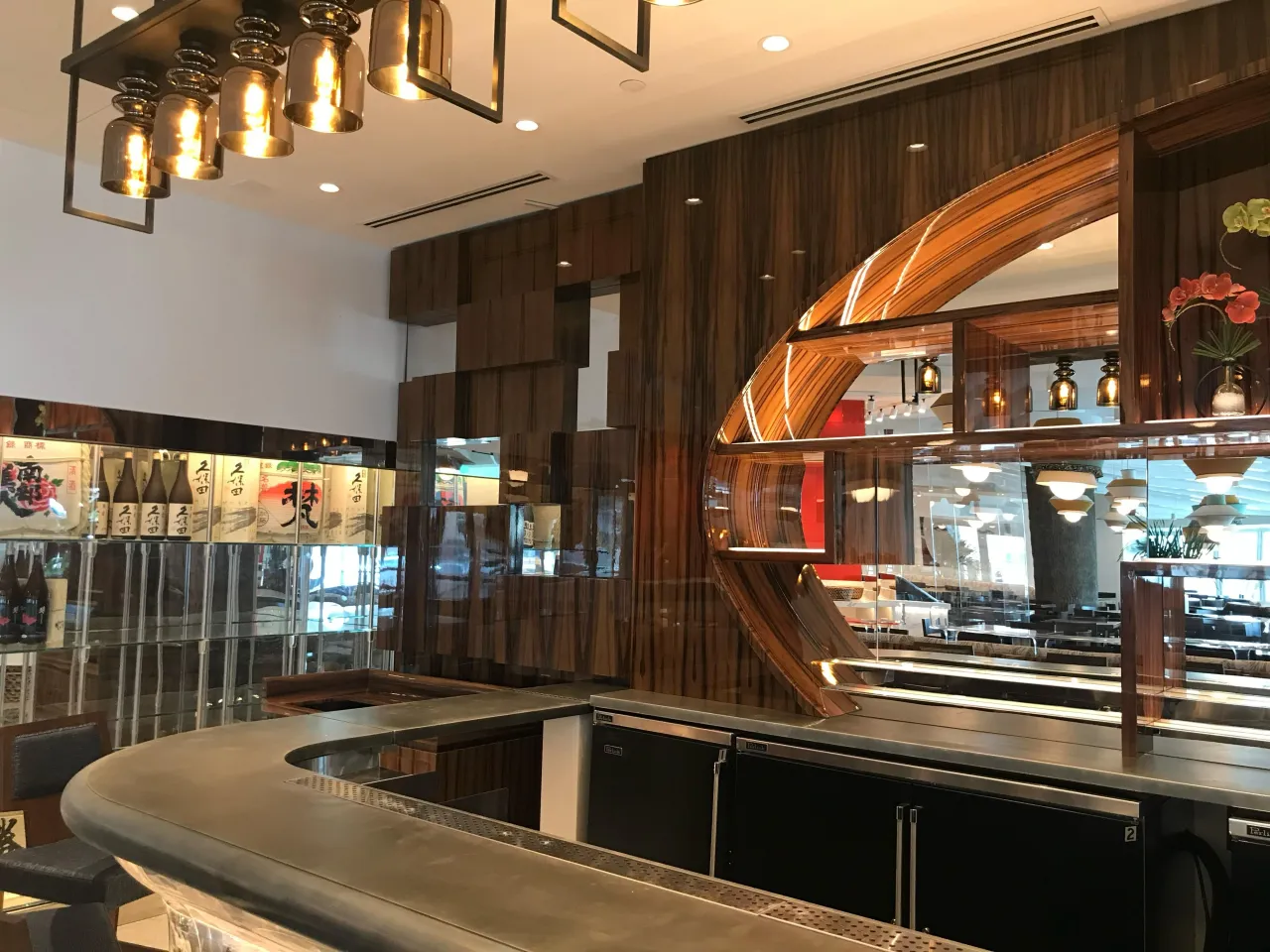 Service bar with a metallic counter curving at the corner, dark refrigerated units below, an oval wood-grain shelving surround, bottle case at left