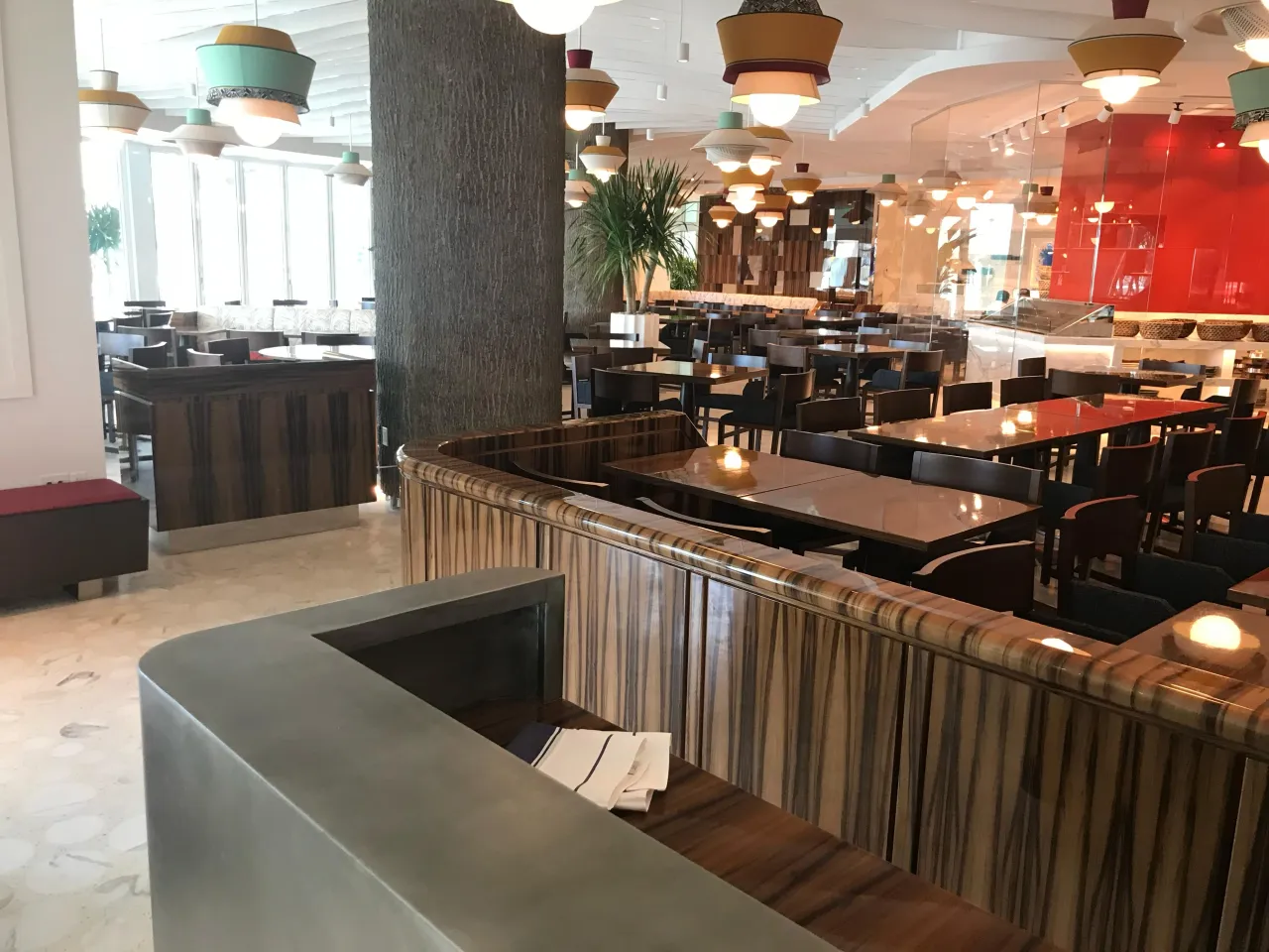 Service station with a metallic-topped counter, curved wood-grain paneled divider behind it, dining tables and colorful pendant lights beyond