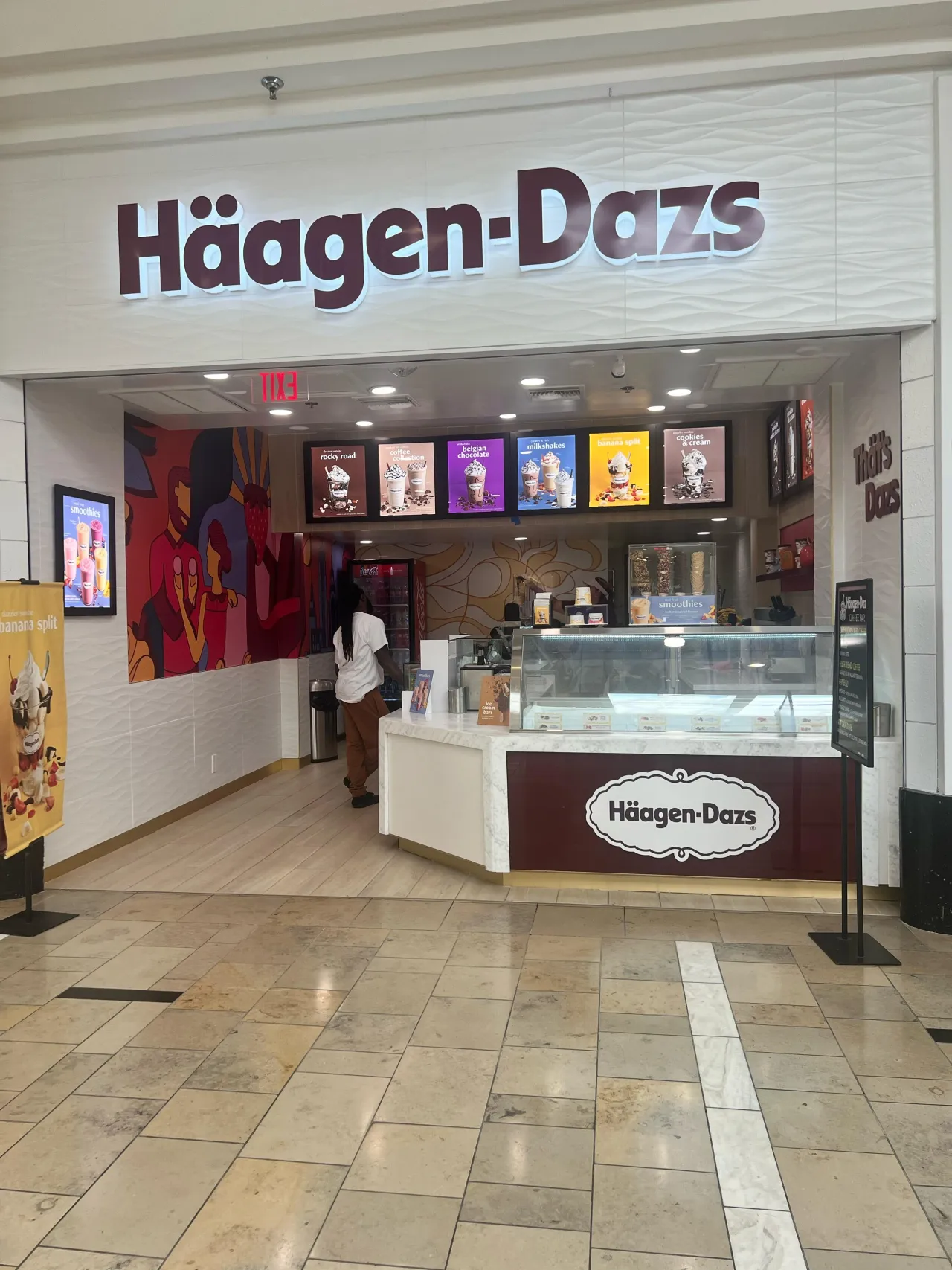 Storefront entrance with a large illuminated sign, wave-textured wall, colorful mural, veined stone counter with maroon branding, mall tile floor