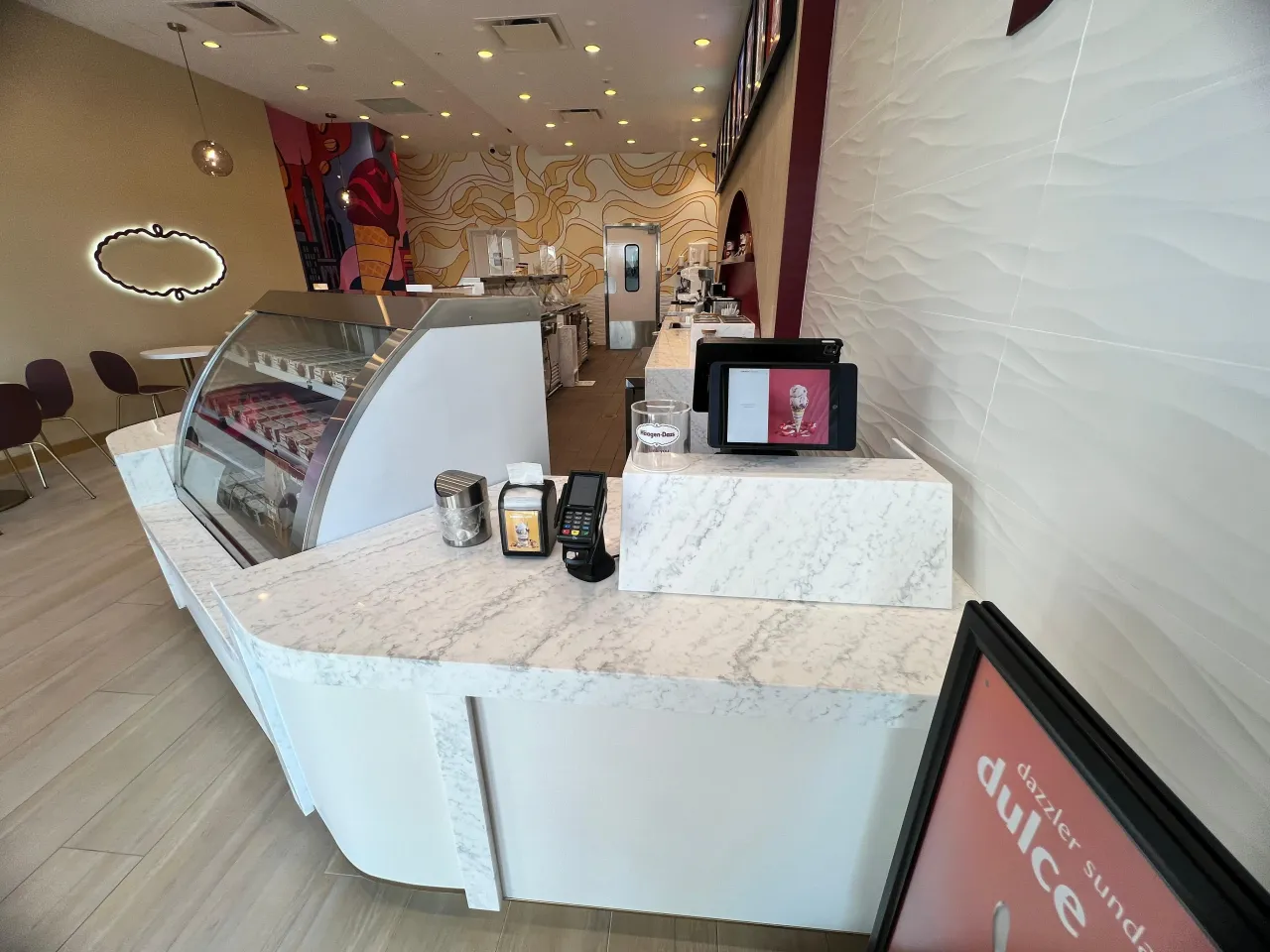 White veined stone checkout counter with curved front, POS tablet and card reader, textured wave wall panel behind, sundae menu sign at right