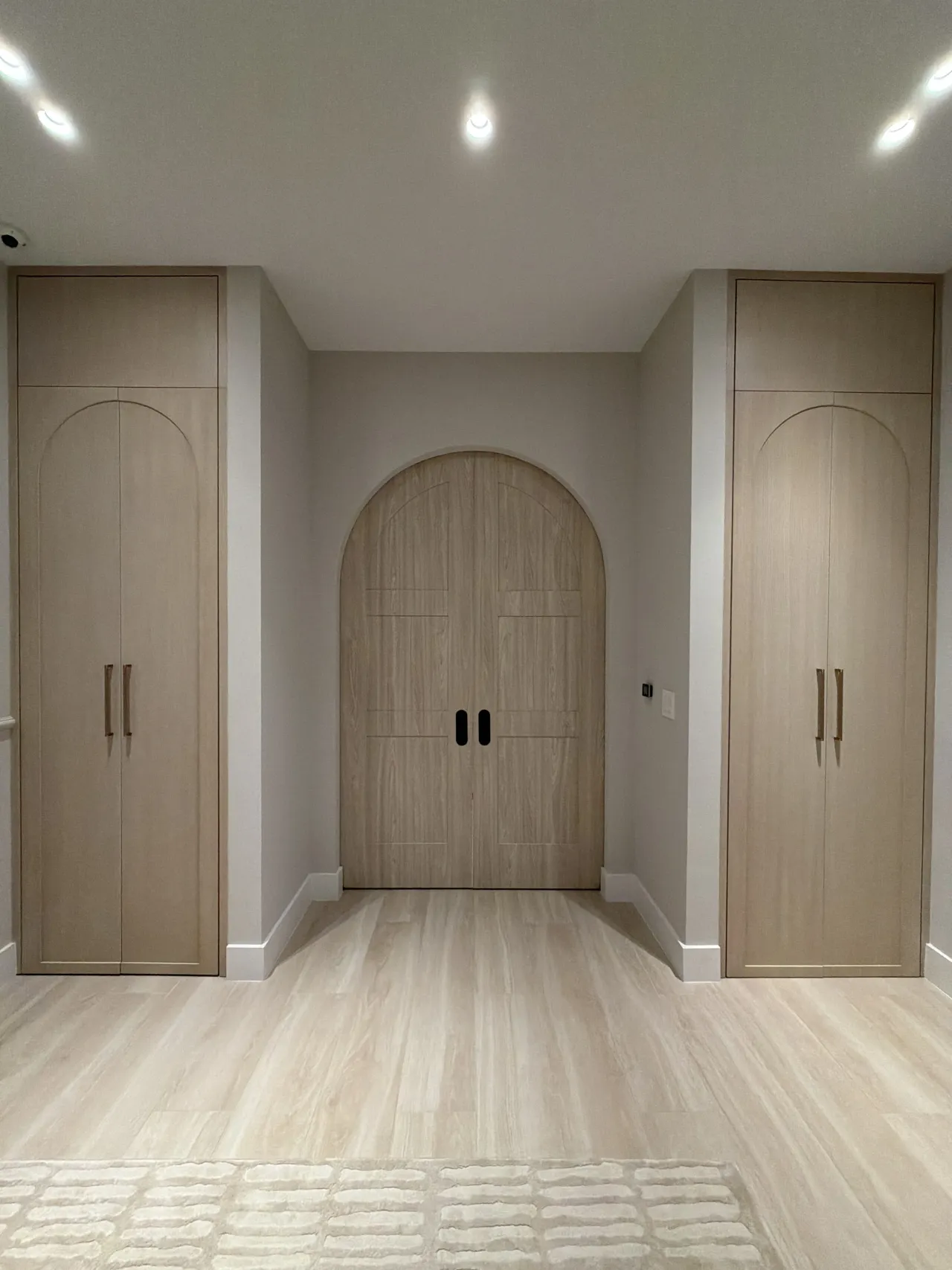 Arched light melamine double doors set in a niche, flanked by two tall built-in wardrobe cabinets with arched inlay detailing and gold-tone pulls