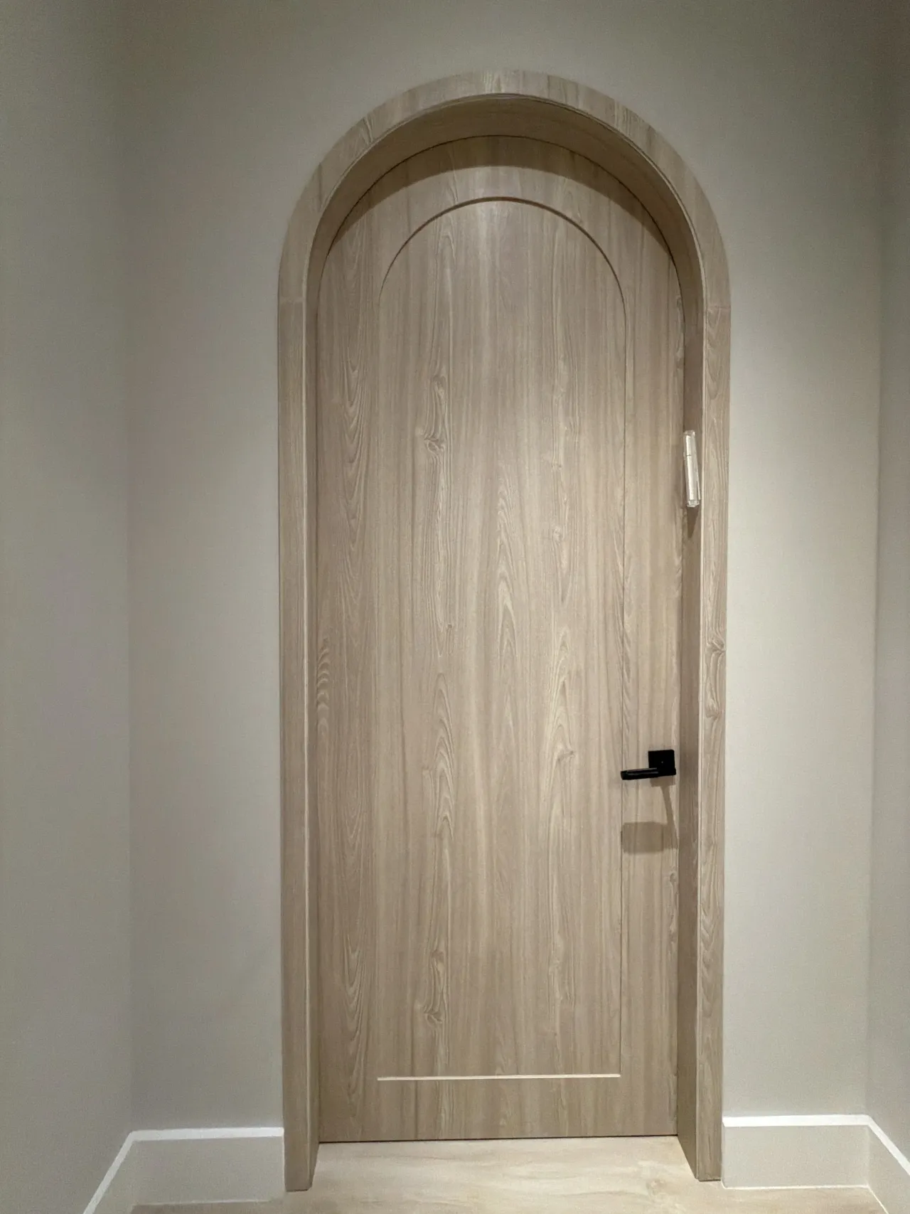 Arched single door in light wood-grain melamine with a full arched wood casing and matte black lever handle, set in a plain light-gray wall