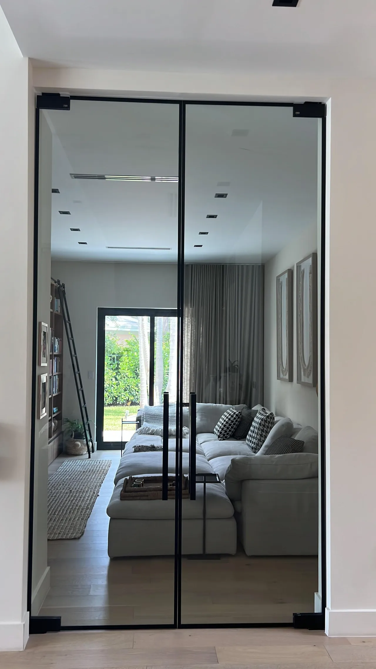 Black-framed glass double doors in a white opening, showing a living area with light sectional sofa, bookshelves with a rolling ladder, wood floor