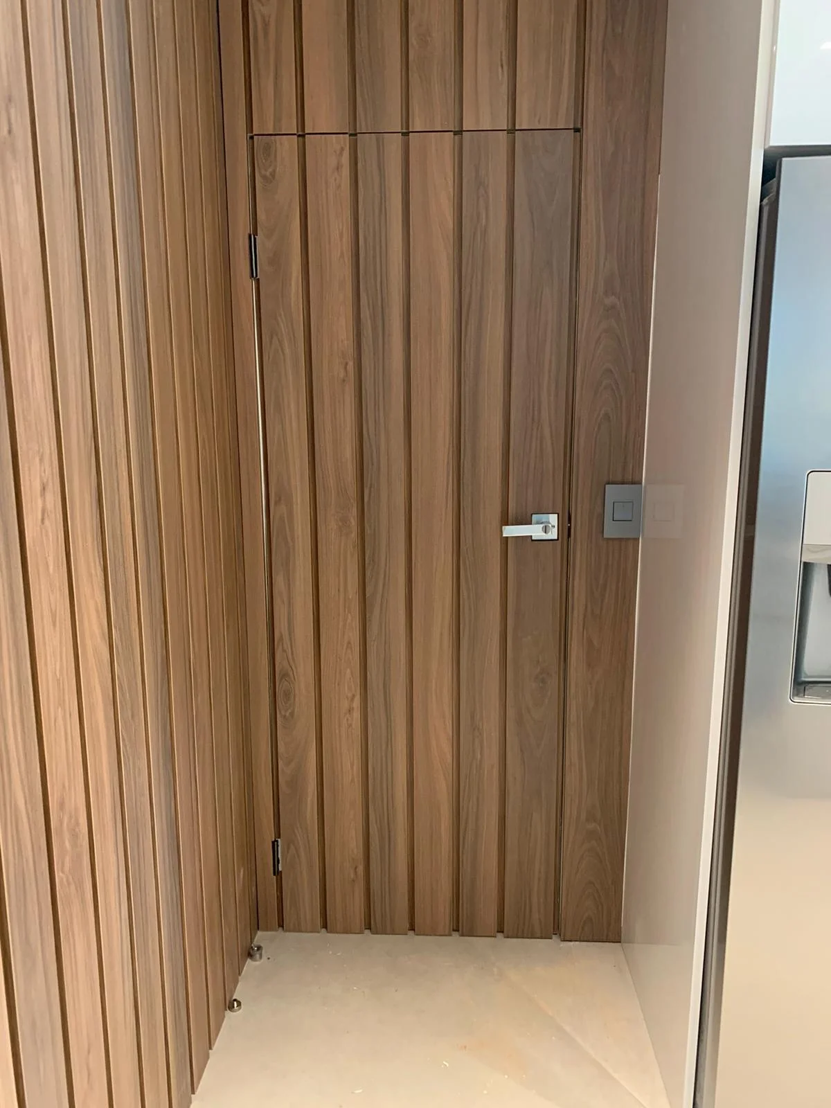 Concealed flush door in a medium wood-grain vertical-slat wall with a slim metallic lever and a square wall plate beside it, light tile floor