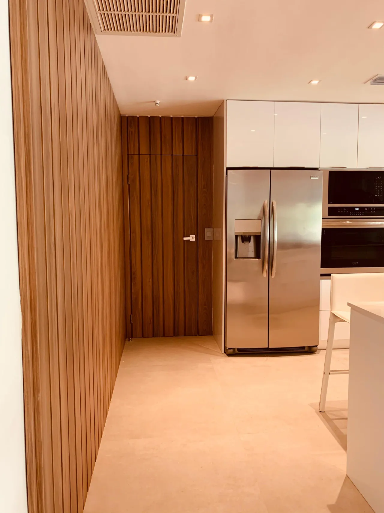 Corridor lined with wood-grain vertical slats ending in a concealed flush door, beside white high-gloss upper cabinets and a stainless refrigerator