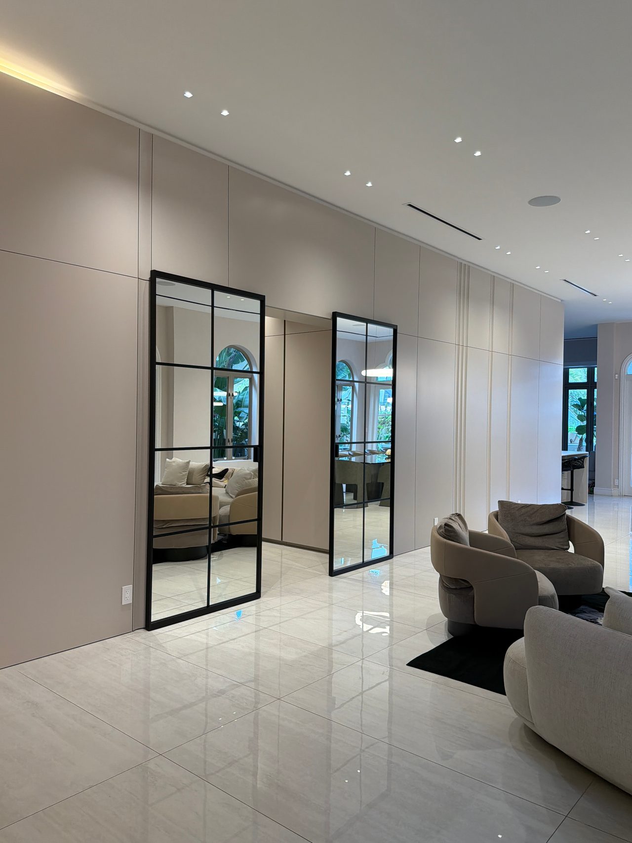 Cream flat-panel wall with two tall black-framed grid-glass doors opening to a living area with upholstered sofas, over a polished stone tile floor