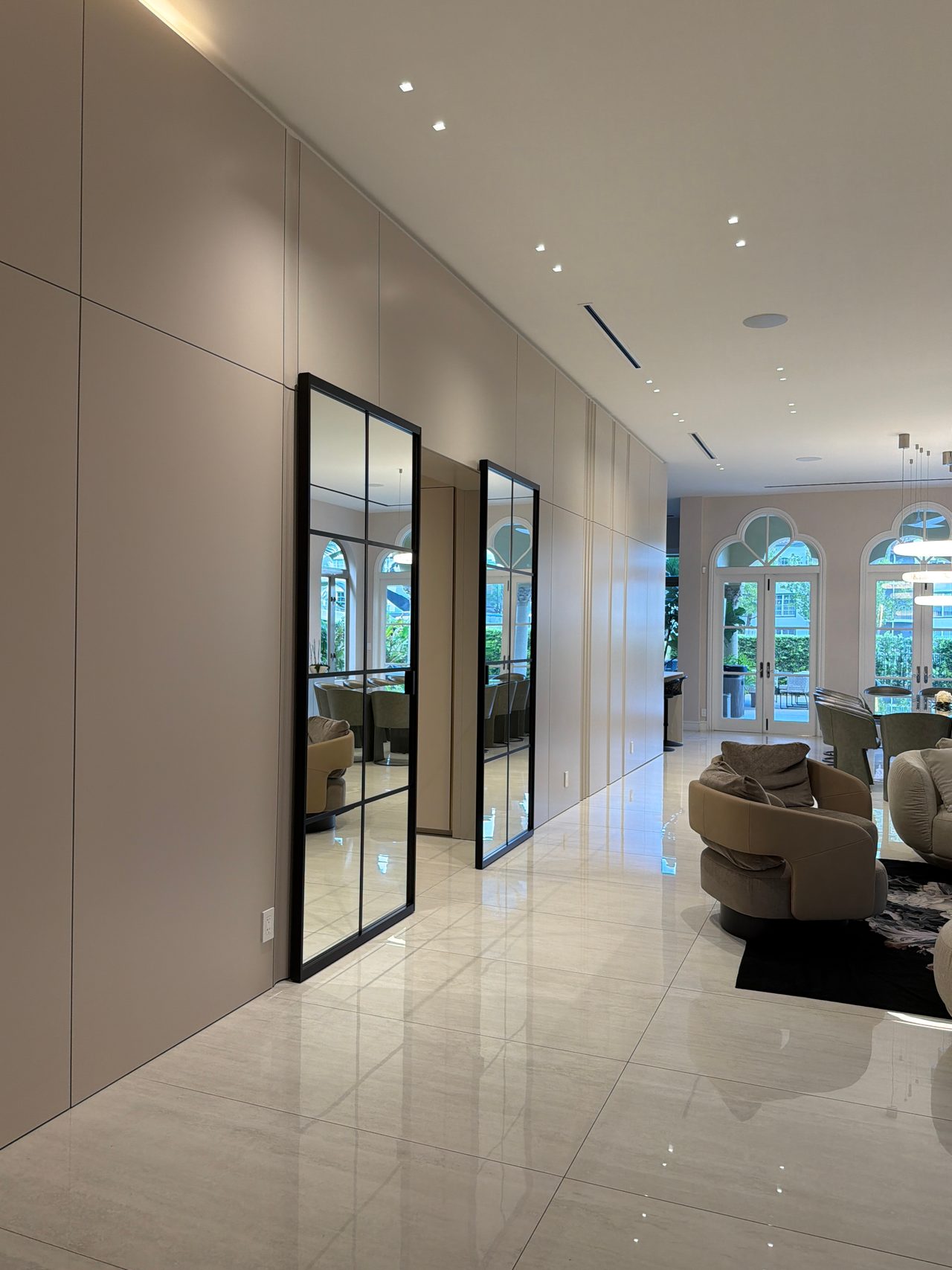 Cream flat-panel wall with two tall black-framed grid-glass doors reflecting arched windows, curved upholstered chairs on polished stone tile