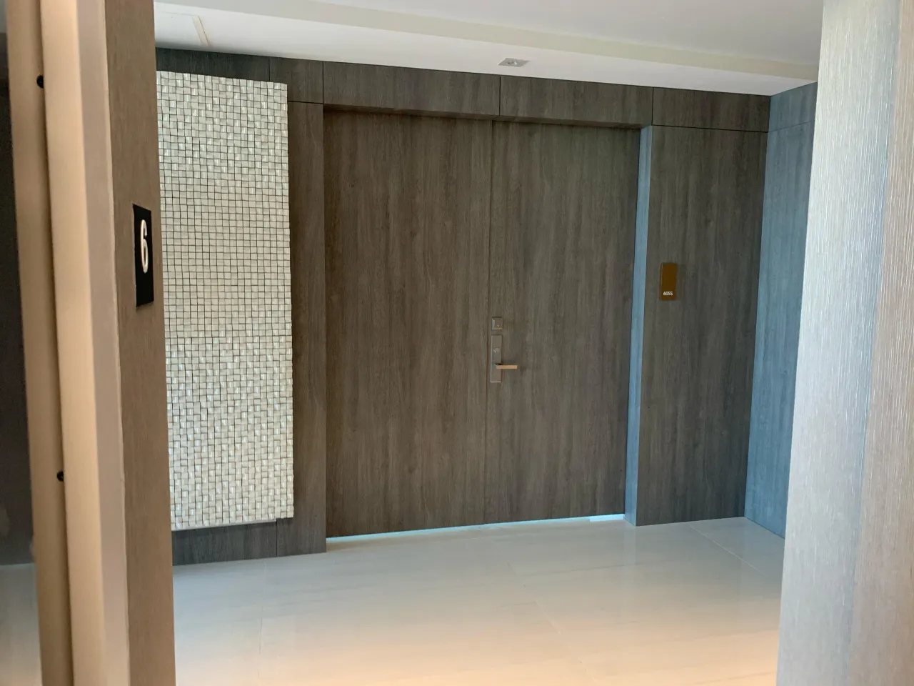 Dark wood-grain double entry door with a gold-tone lever set, in a wood-paneled entry with a white mosaic tile accent panel and light tile floor