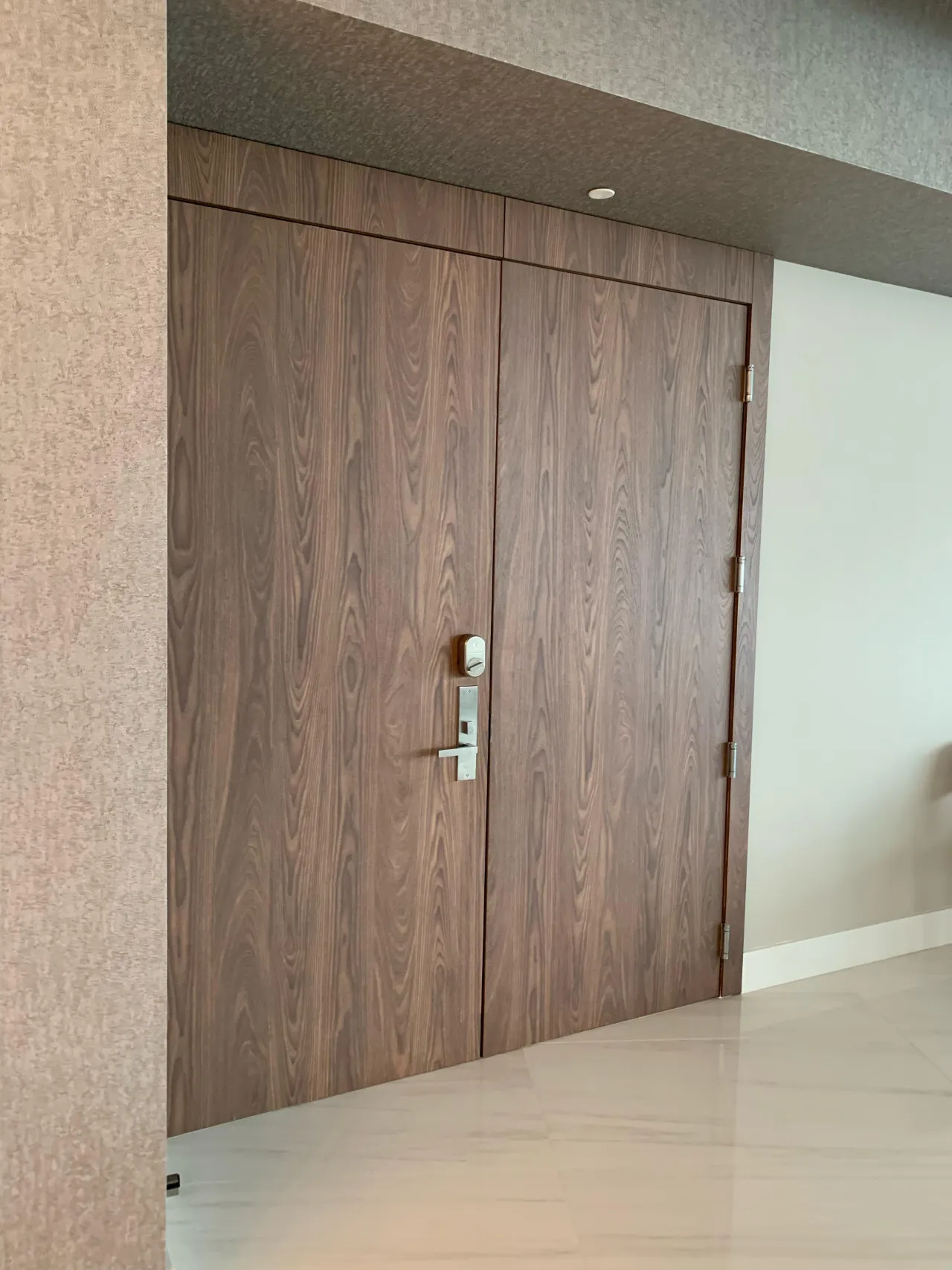 Dark wood-grain flush double entry door with a metallic lever handle and lock plate, set in a textured wall covering above light stone-look tile