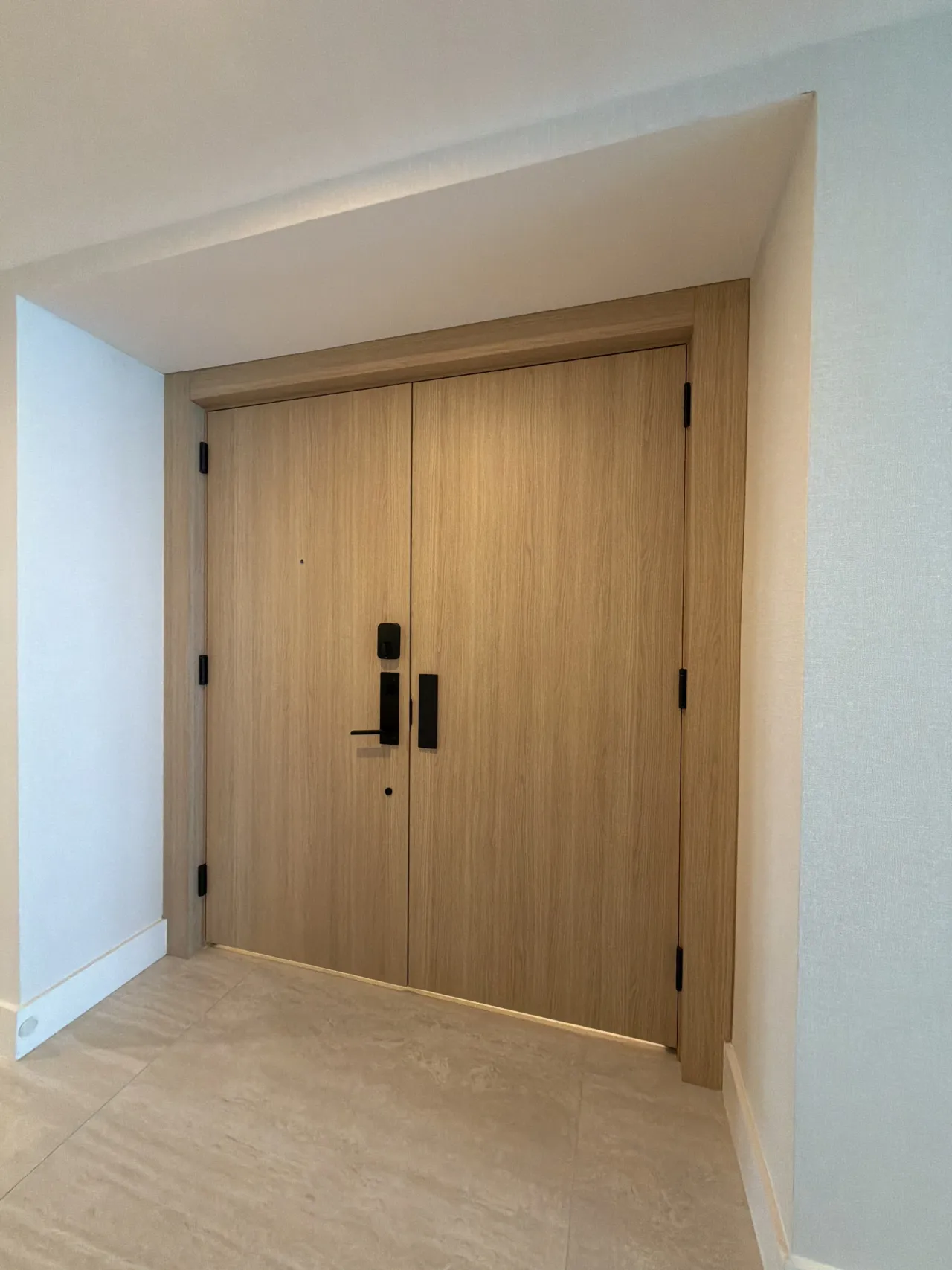 Light wood-grain double entry door with black hinges and a black lever handle, set in matching wood casing, light stone-look tile floor