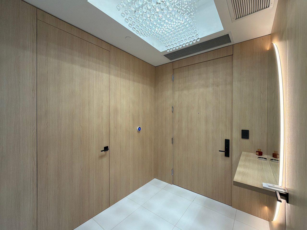 Light wood-grain flush door set into a paneled vestibule wall beside a backlit round mirror and floating shelf, with a black lever handle