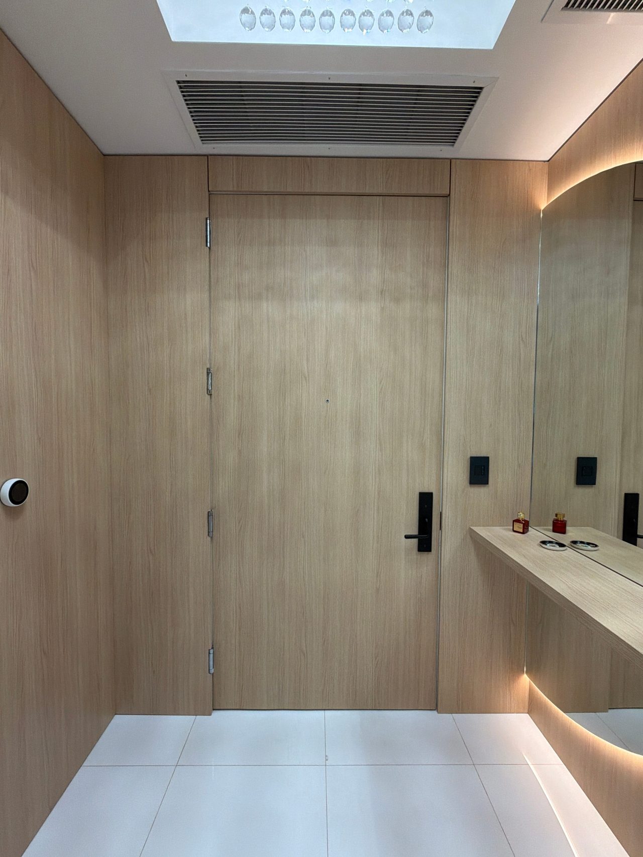 Light wood-grain flush door with a black digital lock and lever, beside a floating shelf and backlit mirror, under a ceiling air grille