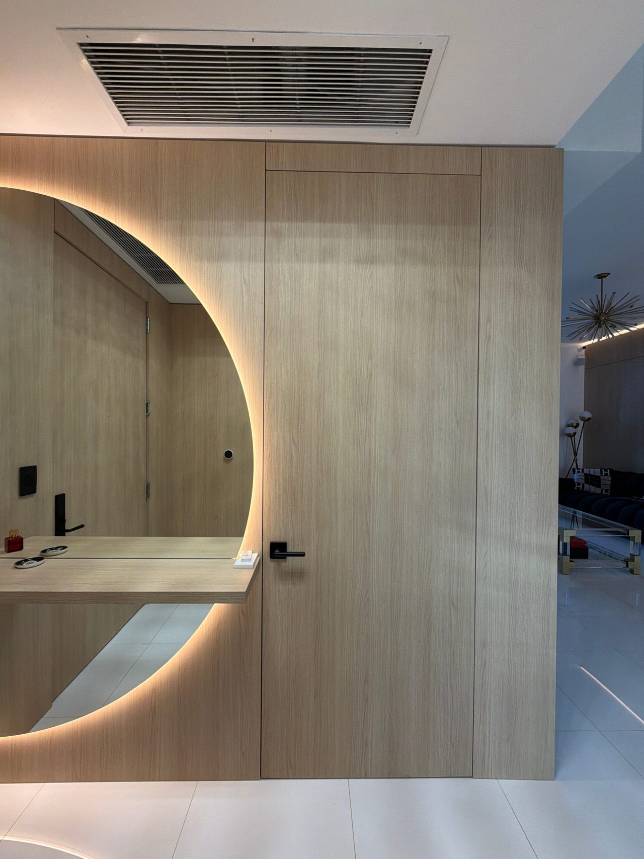 Light wood-grain flush door with a black lever handle beside a half-round backlit mirror and floating shelf, with an air grille above