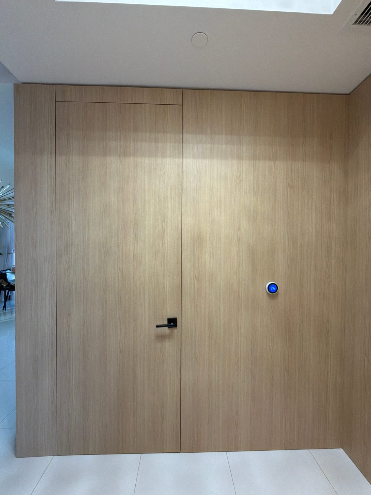 Light wood-grain flush door in a full-height paneled wall with a black lever handle and a wall thermostat beside it